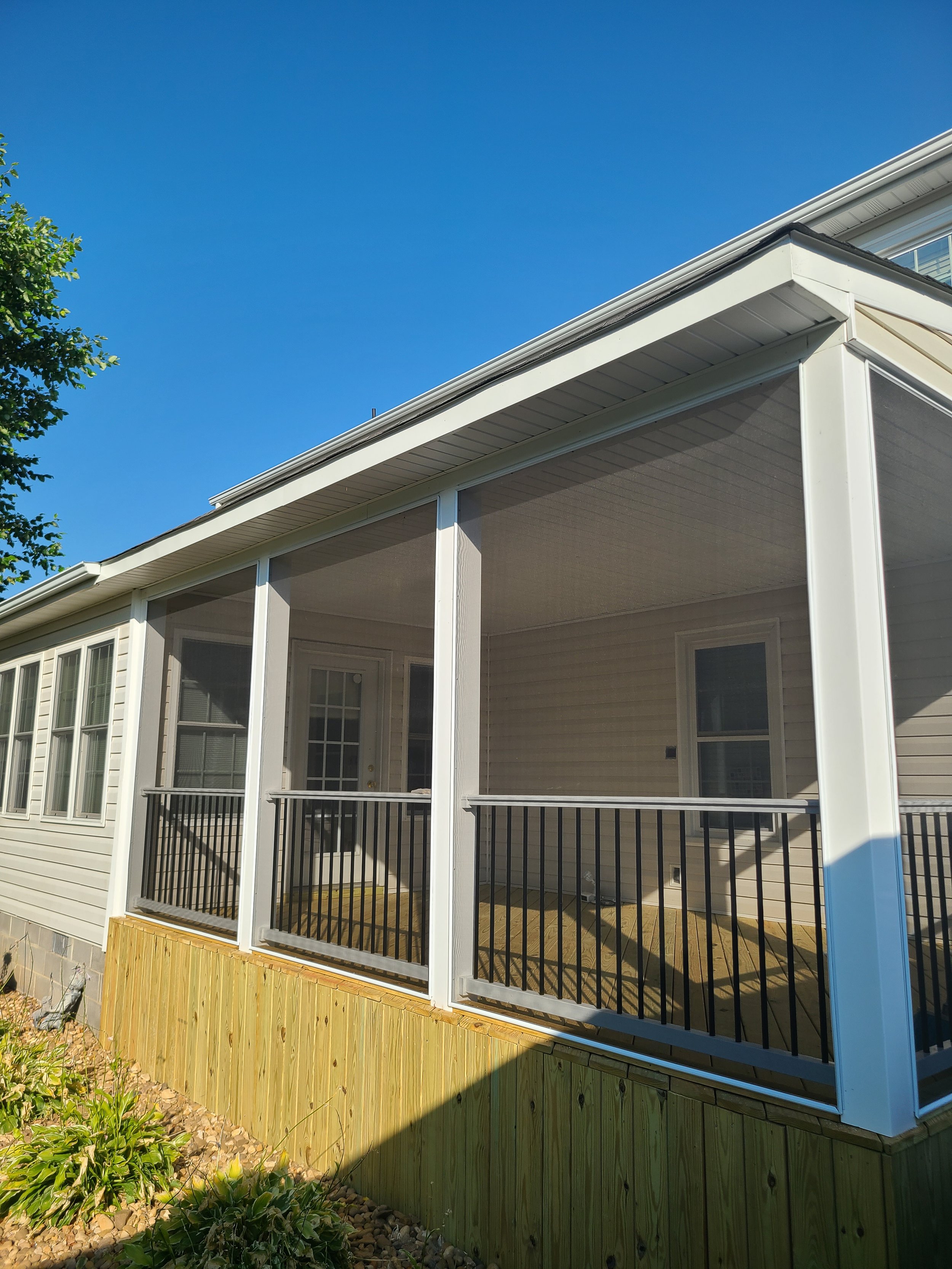 View of a house with a screened-in porch, black railing, and wooden deck, under a clear blue sky with some trees nearby.
