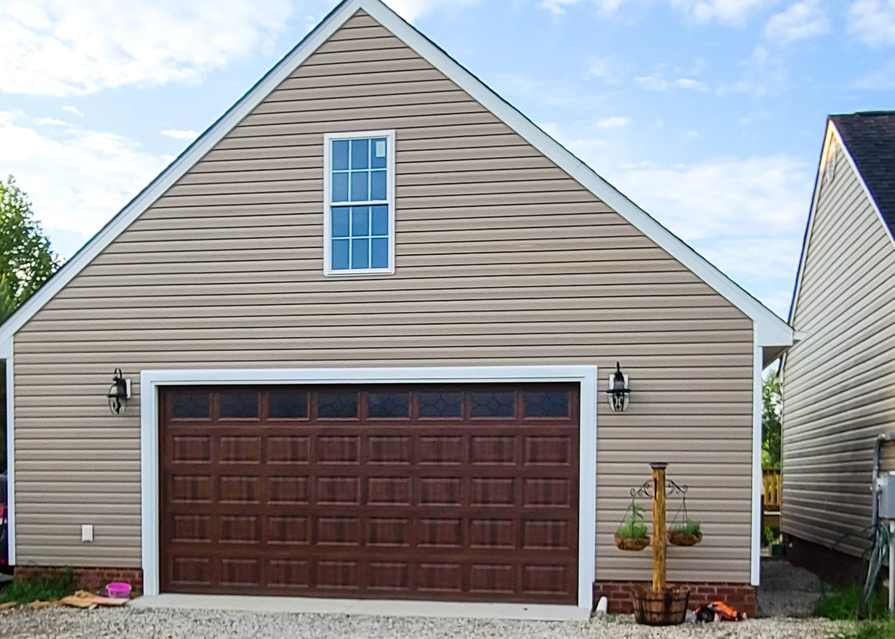 Front view of a beige garage with a brown wooden door, black outdoor lamps on either side, a small window above, and a gravel driveway. There is a hanging plant stand with two baskets to the right and a pink pet bowl on the ground.