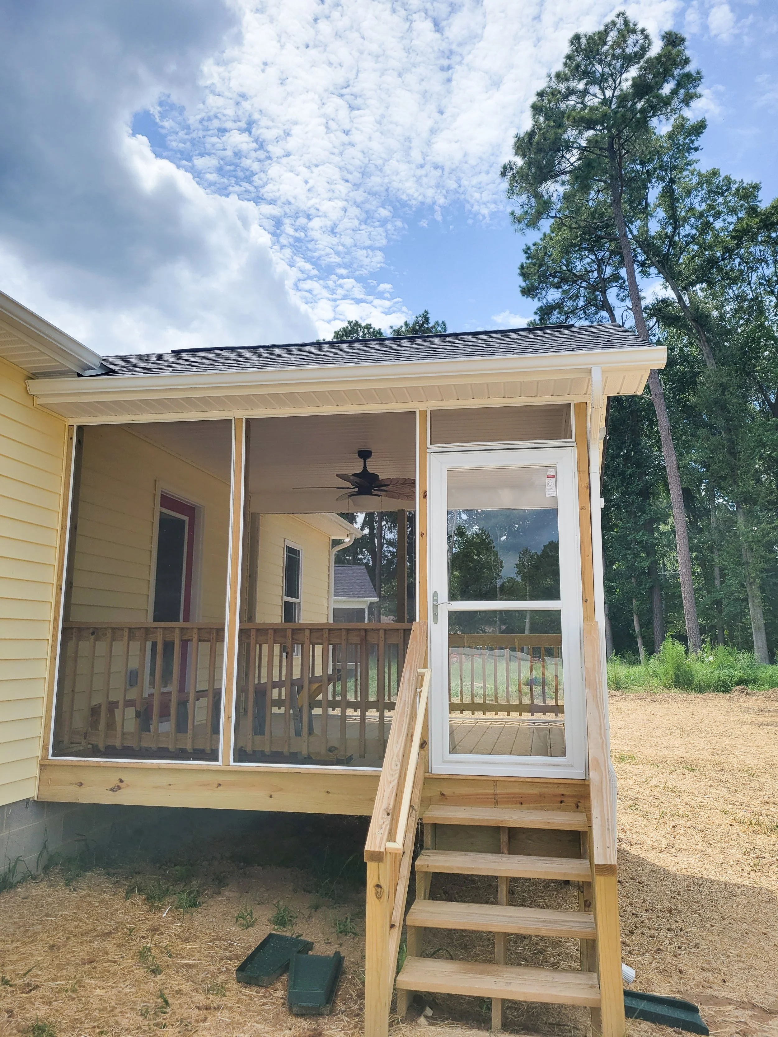 A screened-in porch with a wooden staircase leading up to a glass door, attached to a yellow house, with a blue sky and tall trees in the background.
