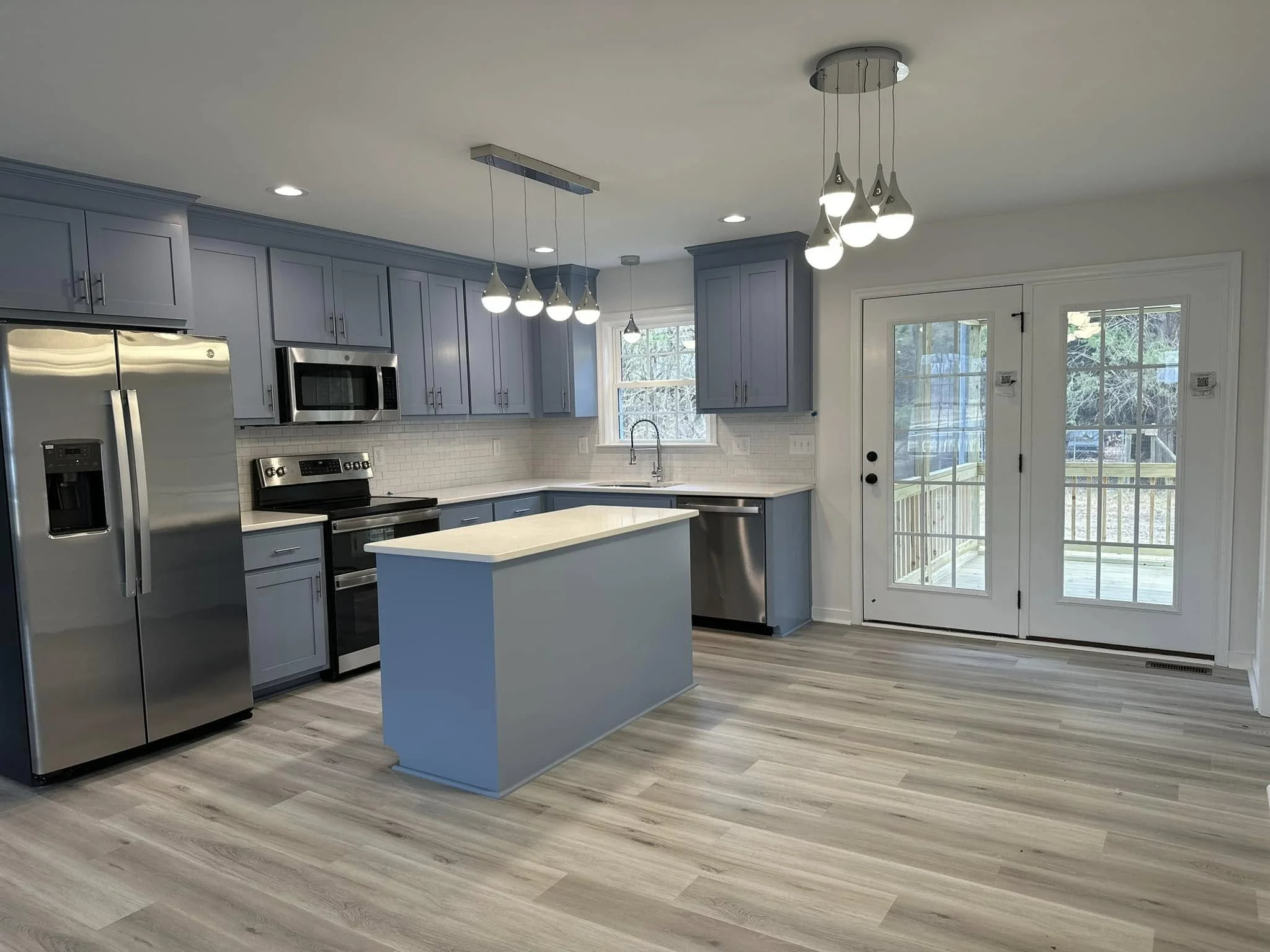 Modern kitchen with blue cabinets, white countertops, stainless steel appliances, and a small island. There are sliding glass doors leading outside, and a window above the sink. Light wood flooring and pendant lights hang from the ceiling.