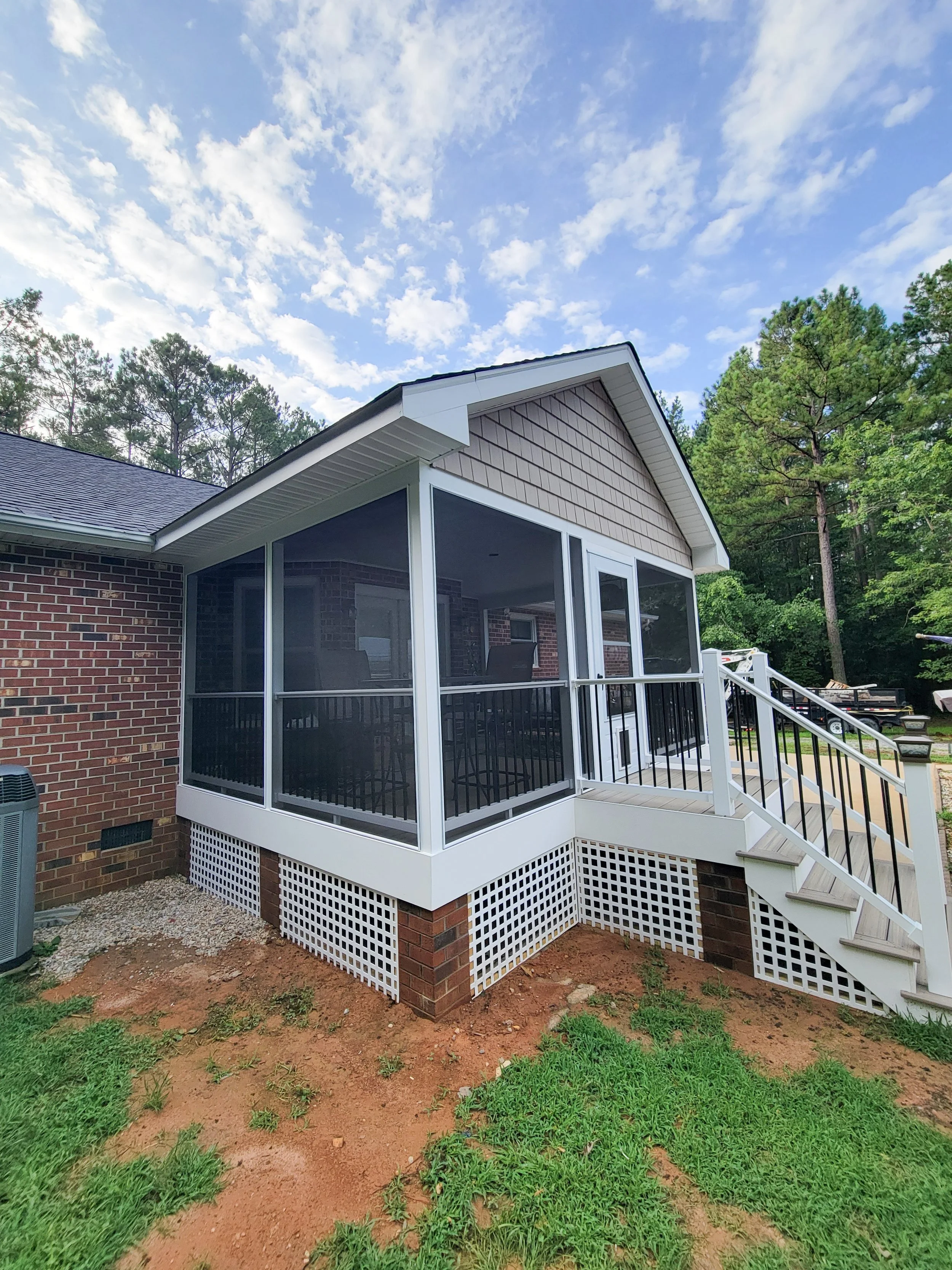 A house with a screened porch elevated on brick and lattice foundation, with steps leading up to it, surrounded by green grass and trees, under a partly cloudy sky.