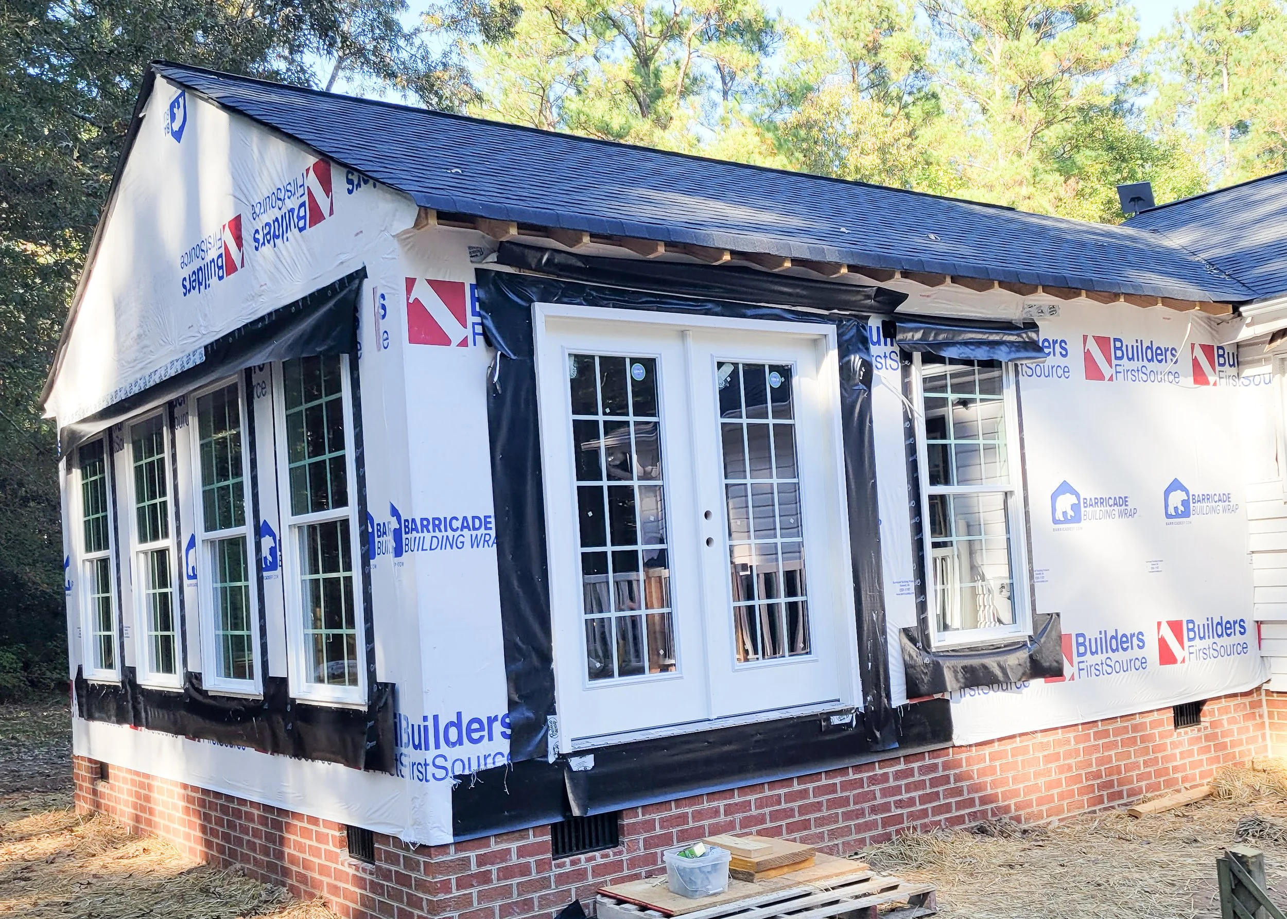 Partial view of a house under construction with black roofing, several white framed windows, a white door with glass panels, brick foundation, and house wrap film with branding, set amidst trees and construction materials on ground.