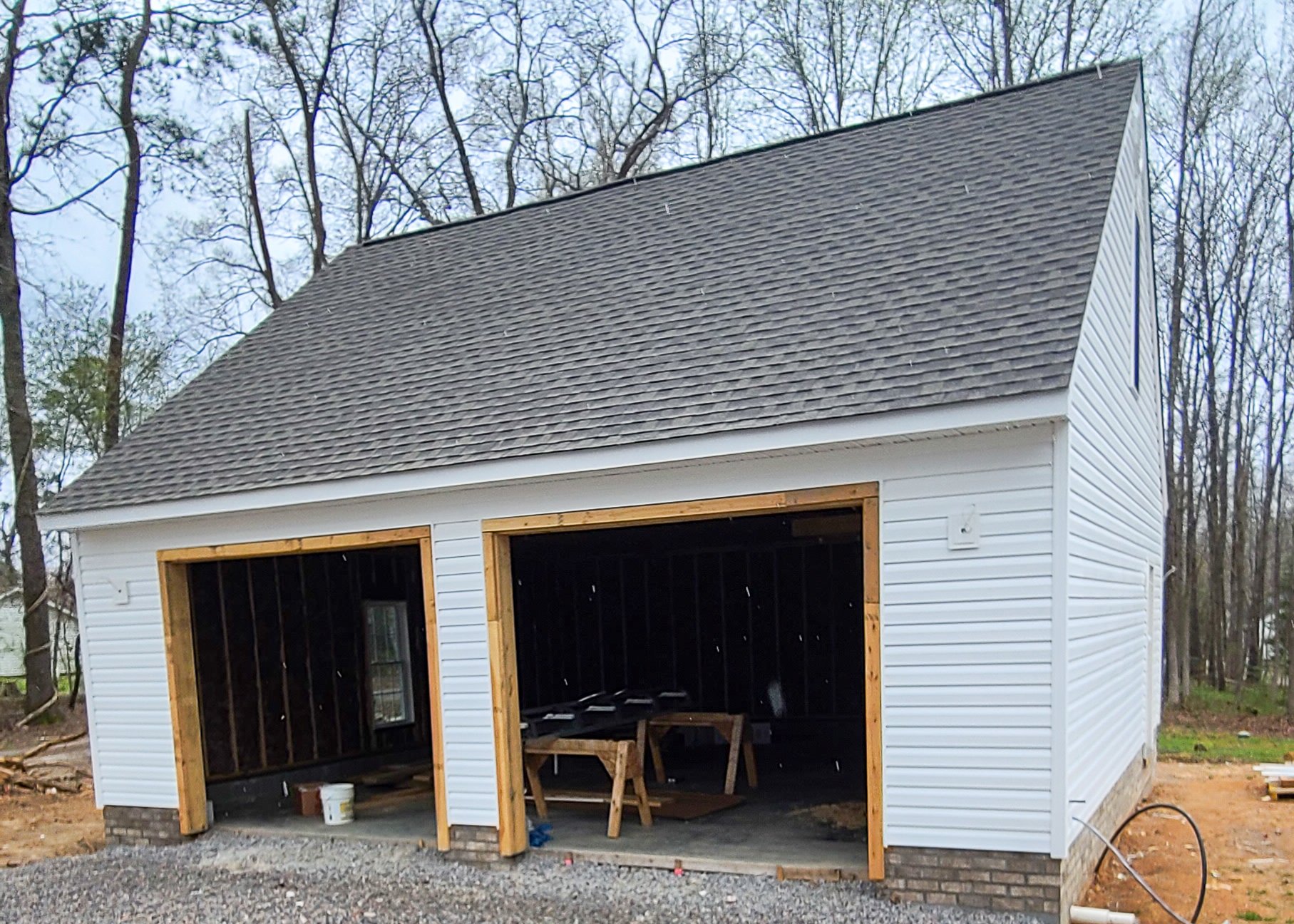 Under construction two-car garage with white siding, black interior, and an asphalt shingle roof, surrounded by leafless trees.