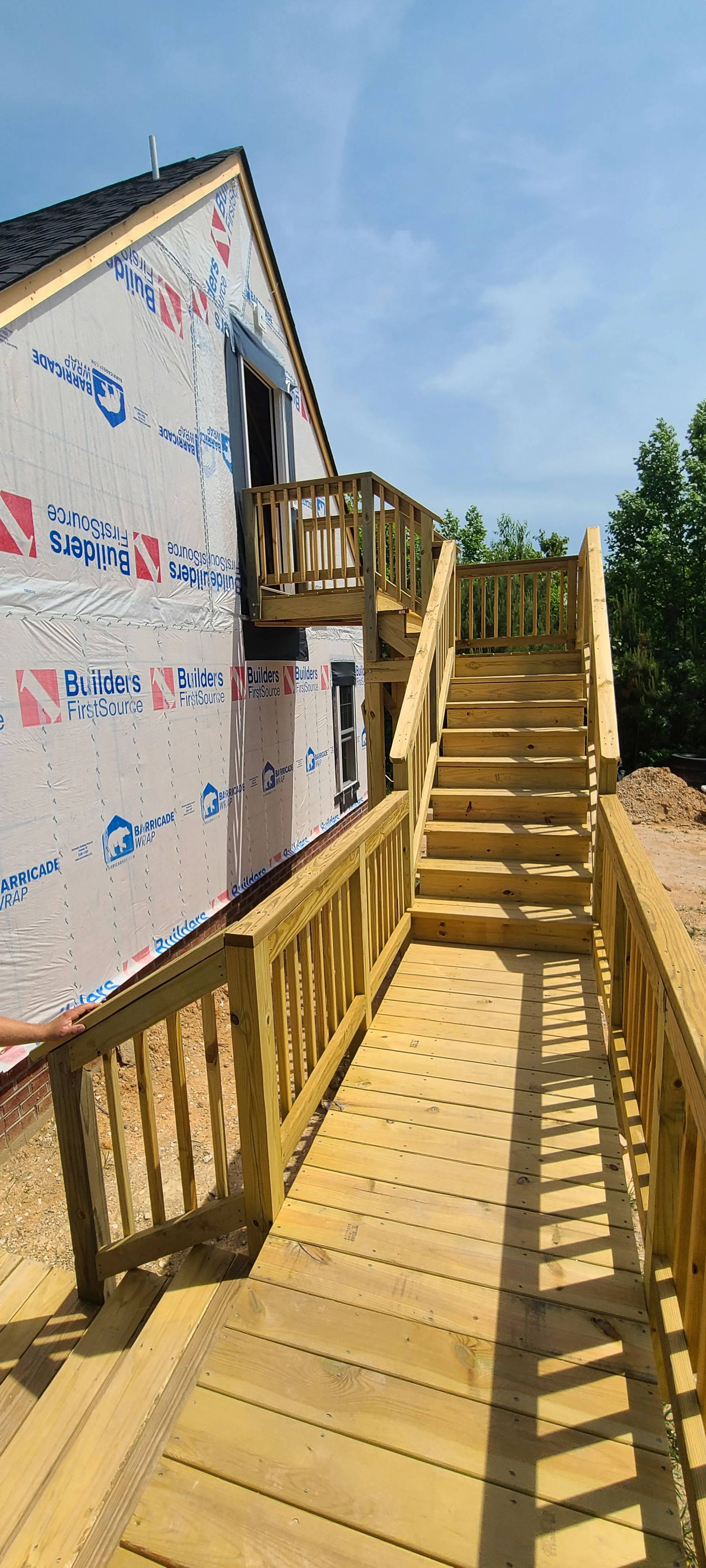 Wooden stairs and ramp outside a house under construction, with an incomplete exterior wall covered in builder's wrap and a small balcony at the top of the stairs under a blue sky.