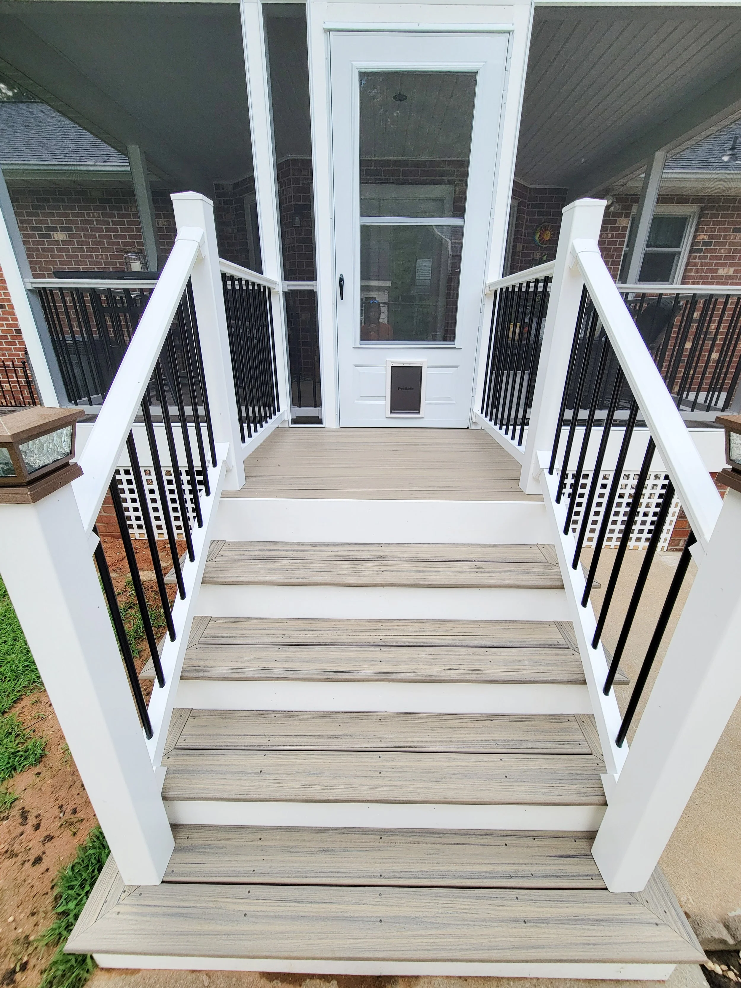 Front porch deck with white railing, beige wood steps, leading up to a screened-in door with a pet door at the bottom, attached to a brick house.