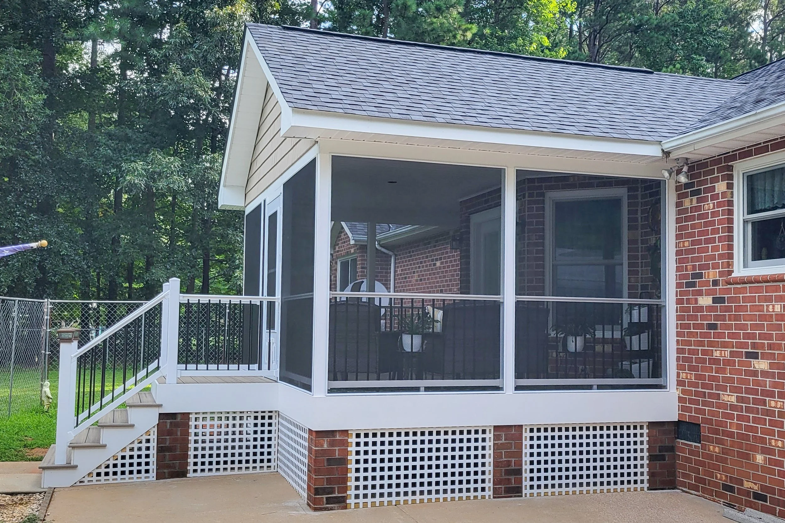 A screened porch attached to a brick house with stairs leading up to it, surrounded by a backyard with a fence and trees in the background.