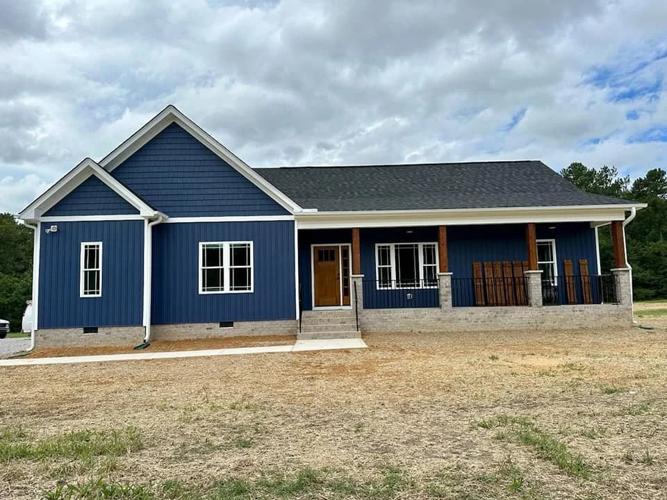 A new house painted in navy blue with white trim, featuring a front porch with wooden columns and black railings, and a small set of stairs leading to the front door. The house has a gable roof and several windows, situated on a dirt lot with a cloud