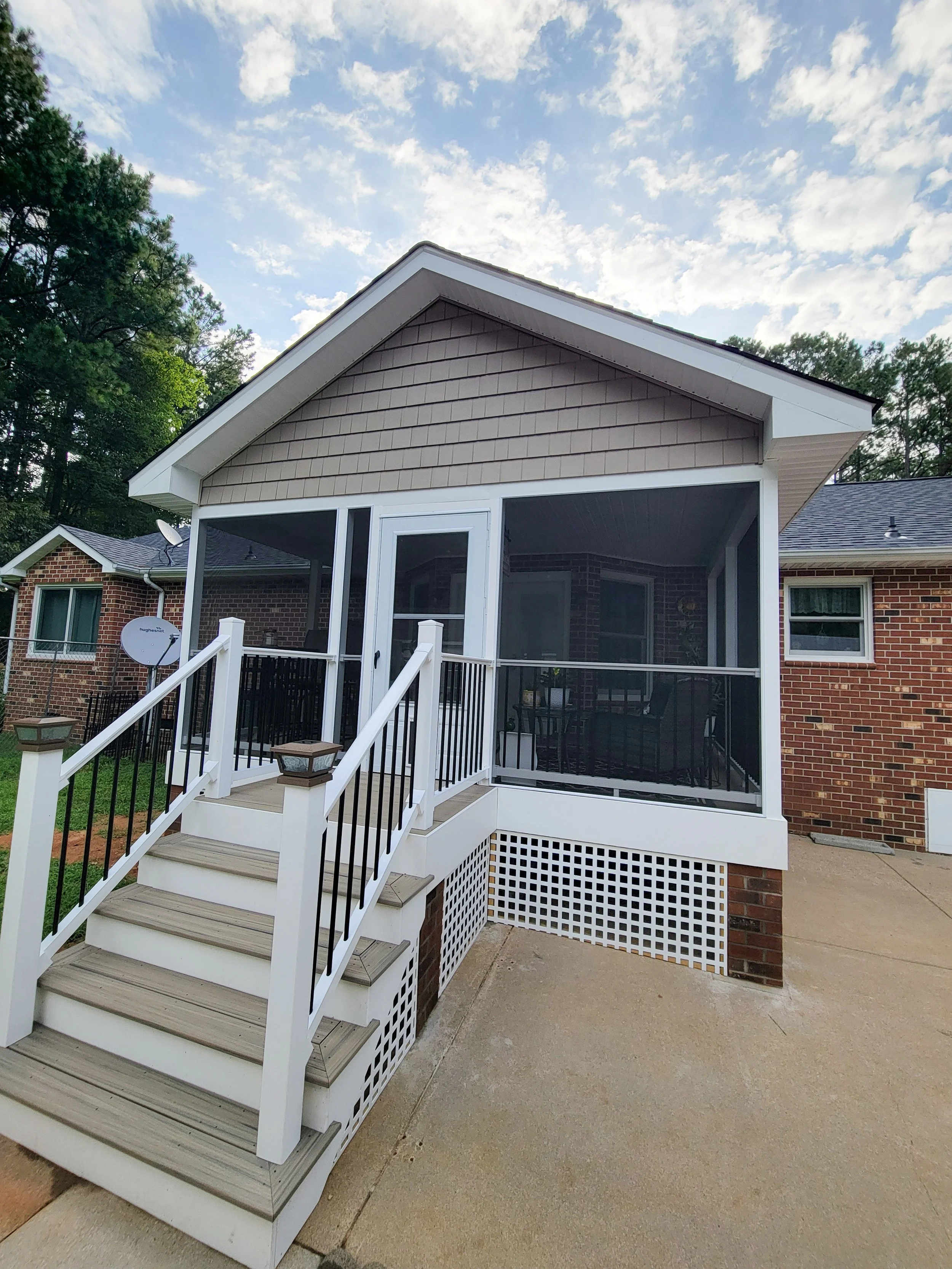 A house with a screened porch, white stairs with black railings, brick foundation, and beige siding under a cloudy sky.