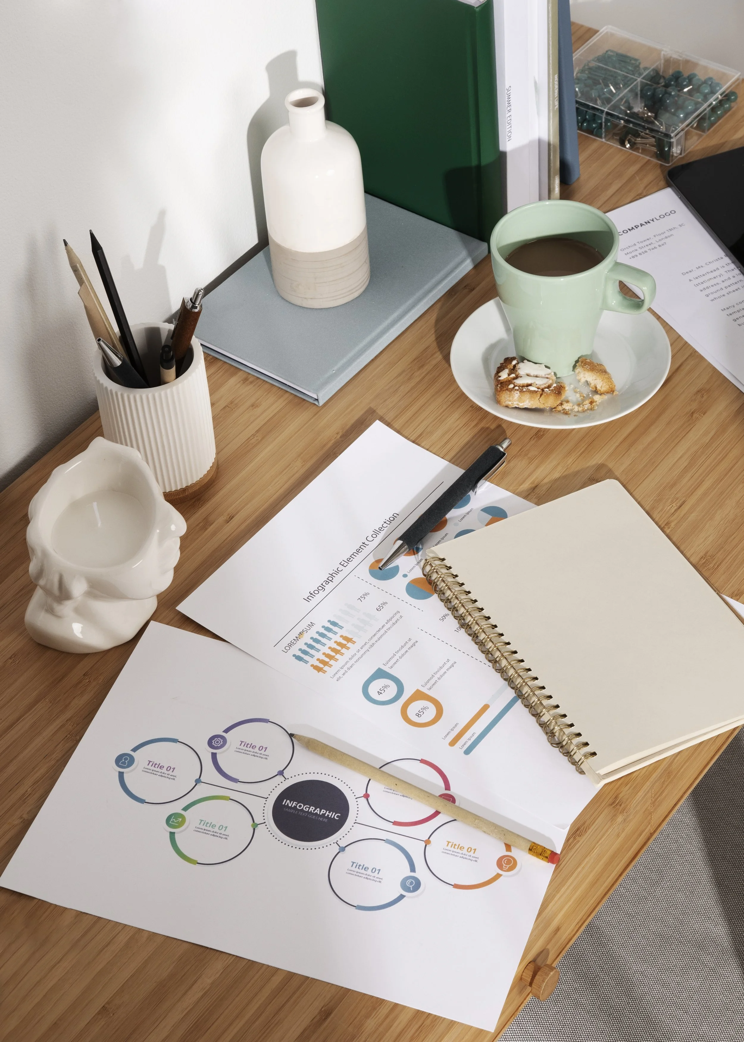 Top view of a cluttered wooden desk with various office supplies including printed infographics, a blank notepad, a black pen, a ceramic head-shaped candle holder, a white striped pen holder with pens, a green book, a white decorative vase, and a cup of coffee with some cookies on a small plate.