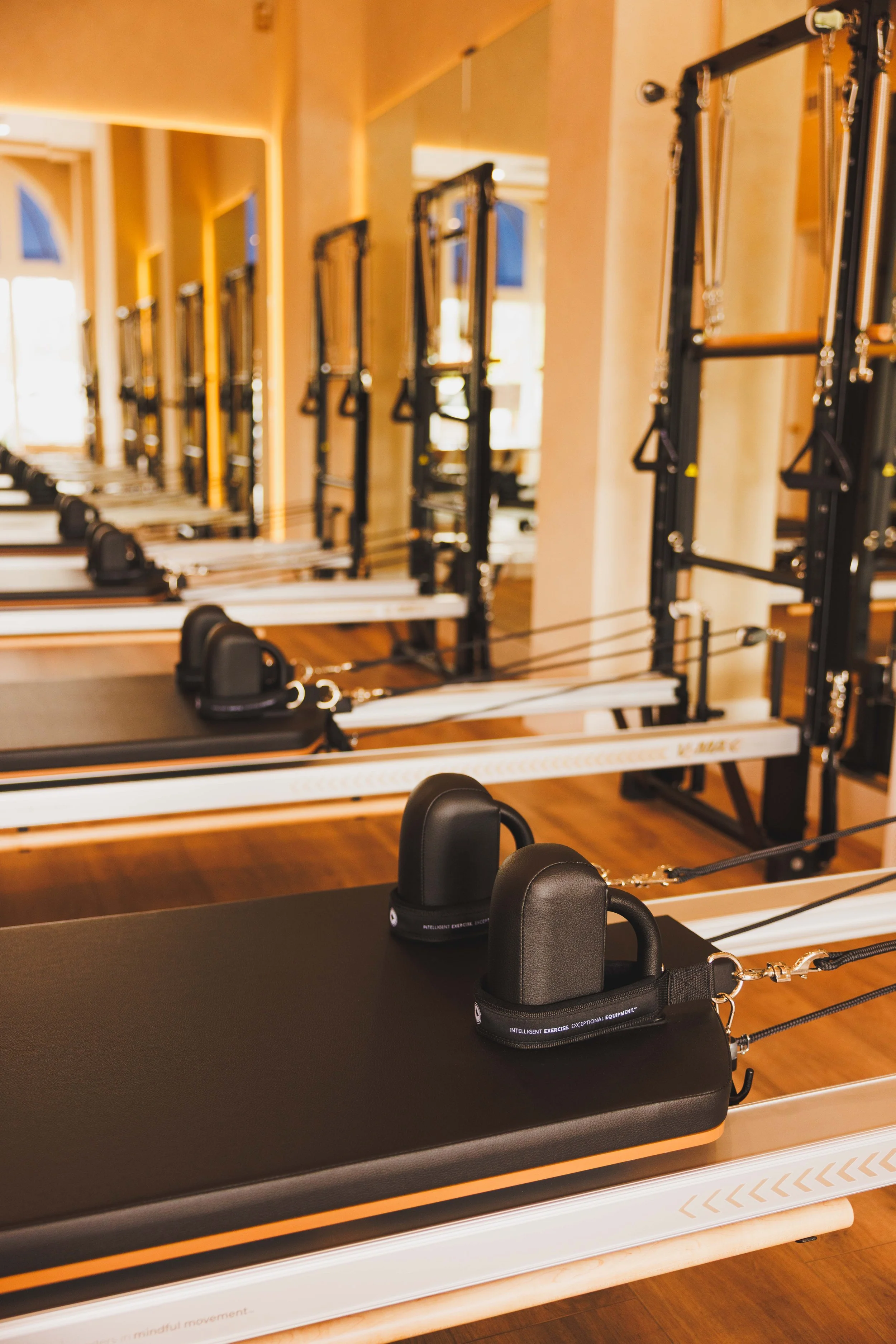 Multiple reformer Pilates machines lined up in a fitness studio with large windows and warm lighting.