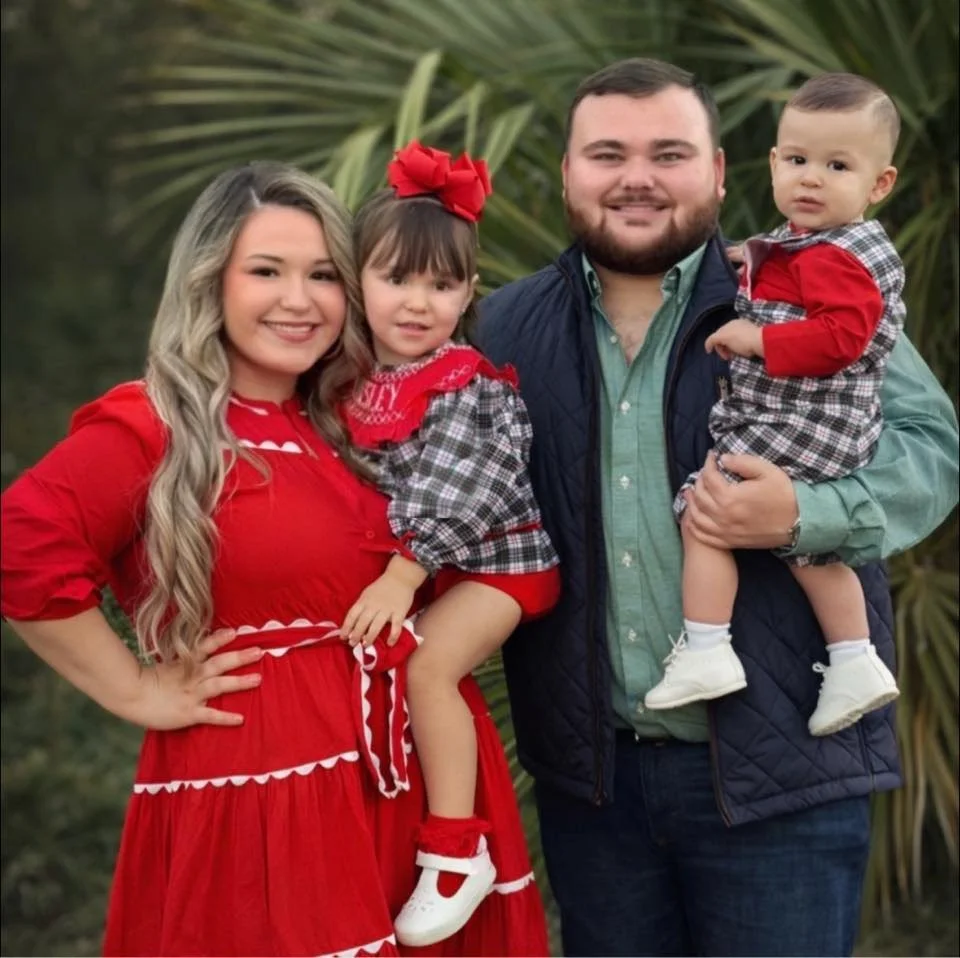 A family of four posing outdoors in front of green foliage, with the mother and daughter dressed in red, and the father and son wearing green and plaid outfits.