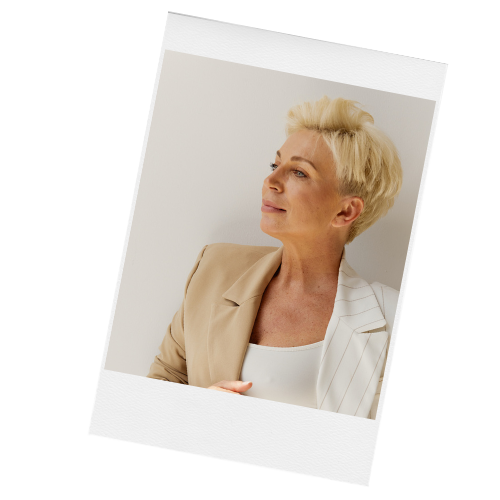 Portrait of a mature woman with short blonde hair, wearing a beige blazer and white top, looking to the side against a plain background.