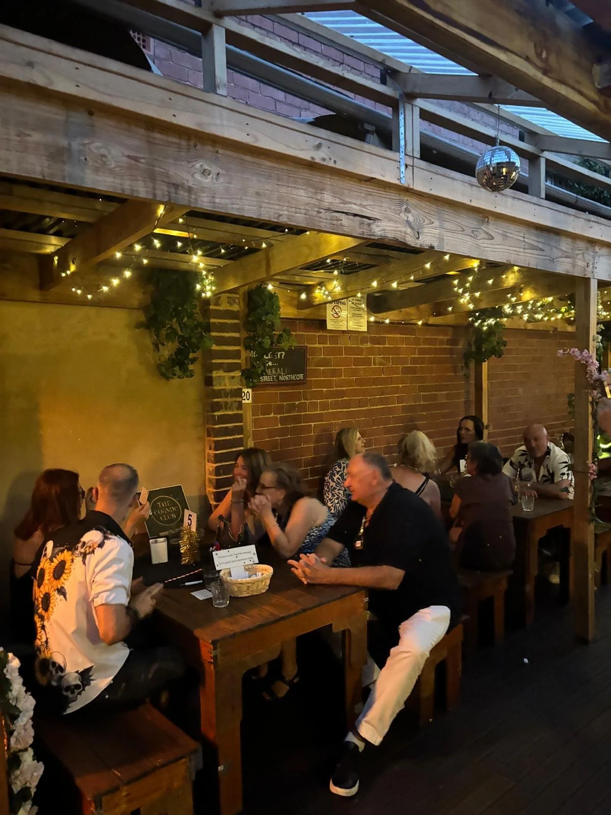 People socializing at a bar or cafe with large windows and hanging lights, holding drinks and engaging in conversation.