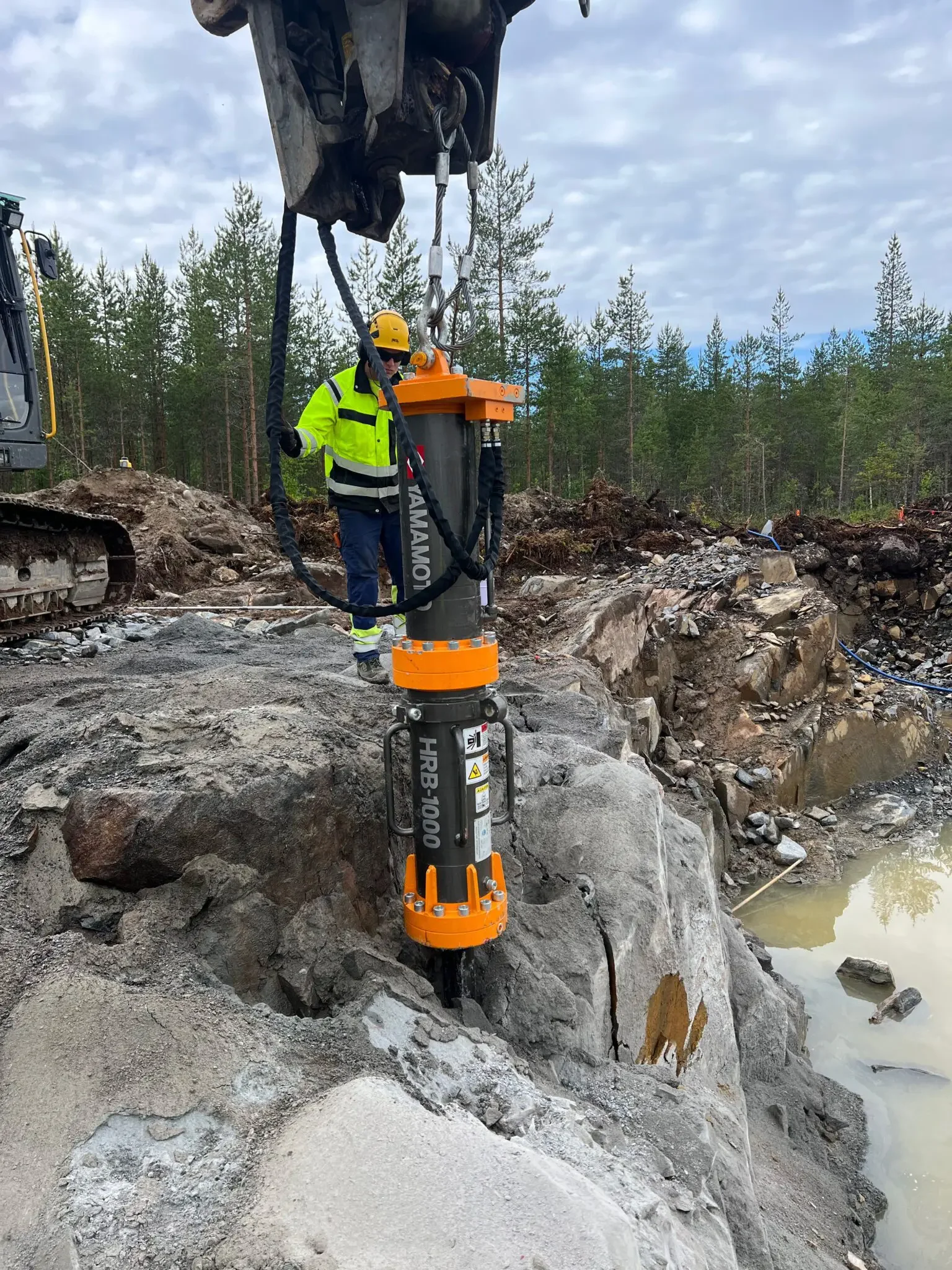 Rakennustyöntekijä työskentelee luonnonmukaisessa ympäristössä korkealla laatan päällä, käyttäen turvakenkiä ja kypärää, ja työskentelee suuren, keltaisen ja mustan työkalun kanssa, jota pidetään pystyssä betonipylväässä.