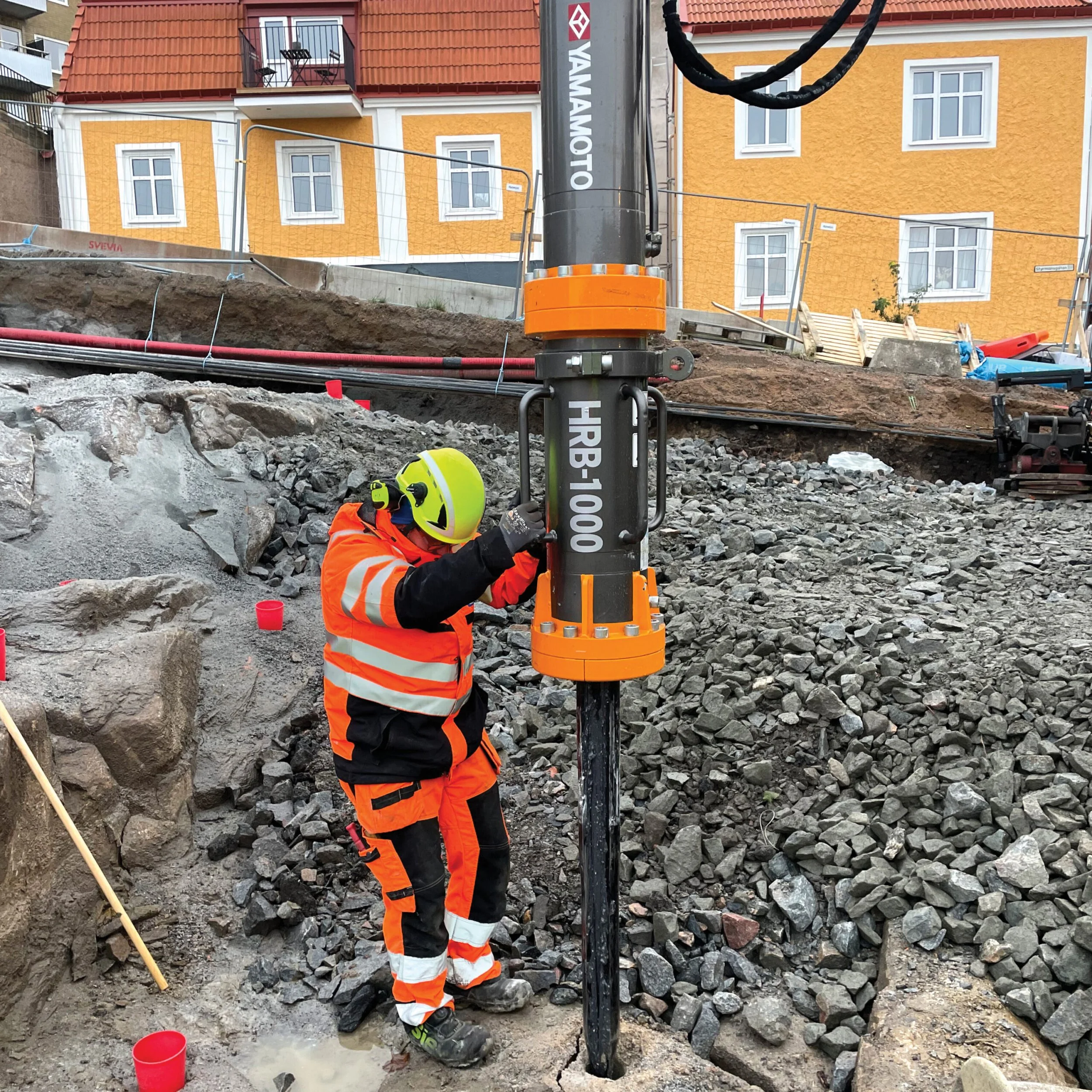 Rakennustyöntekijä pukeutuneena kirkkaan oranssiin ja mustaan työhaalariin, keltainen kypärä ja mustat hanskat, työskentelee kaivannon pohjassa ja kiristää maadoitusta hänen oikeassa kädessään sidottuna pylväseen. Taustalla näkyy kerrostalo ja rakennustelineitä, maassa on kiviä ja rakennustarvikkeita.