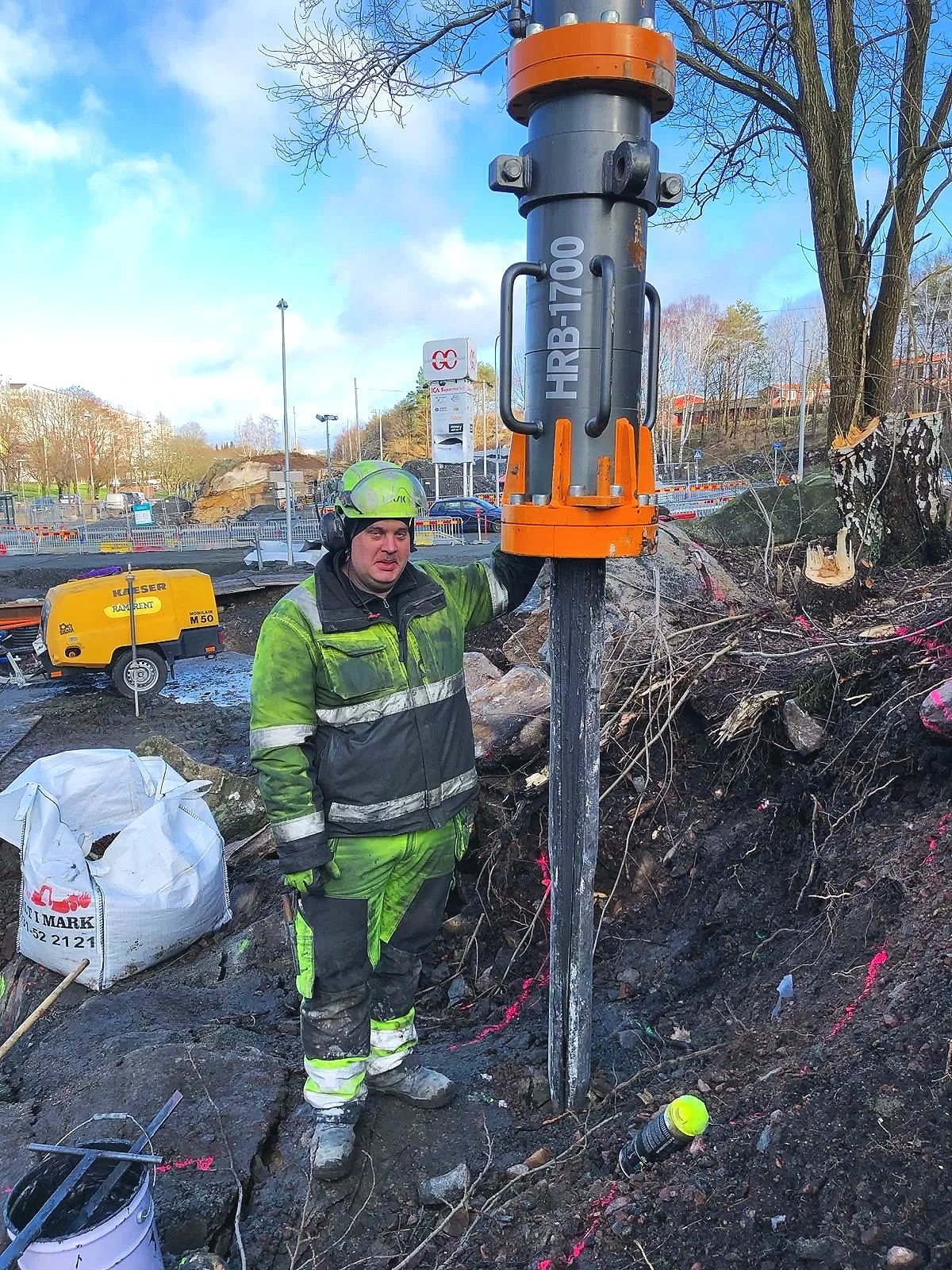 Työmies seisoo maassa, jossa on maaperää ja juuria, ja pitää maahan asennettua porakoneen näppäimistöä. Taustalla on rakennustyömaa ja kalustoa.