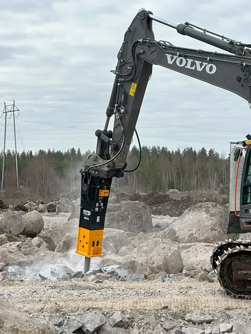Kaivinkone, merkki Volvo, tekee kaivuutöitä kalliolla ja maastossa, ympäristössä puita ja sähkölinjoja.