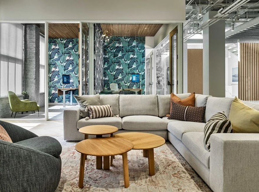 Modern office lounge with a large beige sectional sofa, colorful throw pillows, wooden coffee tables, and a patterned rug. In the background, glass-walled meeting rooms with tropical wallpaper and seating areas. Bright, industrial-style space with large windows and exposed ceilings.