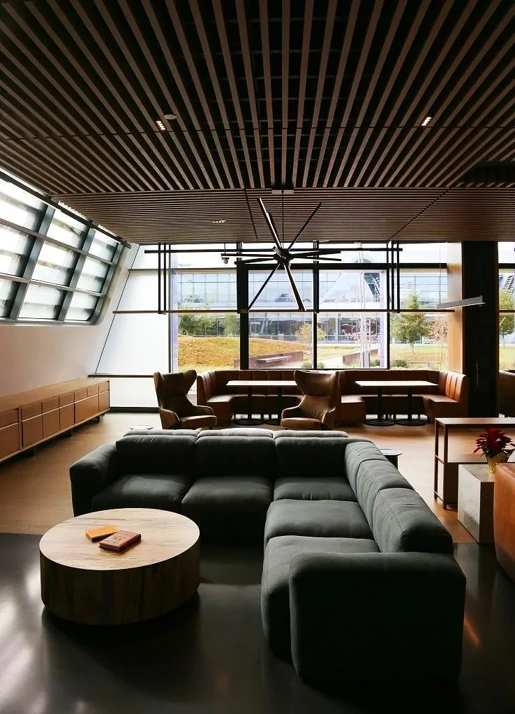 Modern lounge with gray sectional sofa, round wooden coffee table, leather armchairs, large windows, wooden ceiling with linear design, and a large black star-shaped clock hanging from the ceiling.