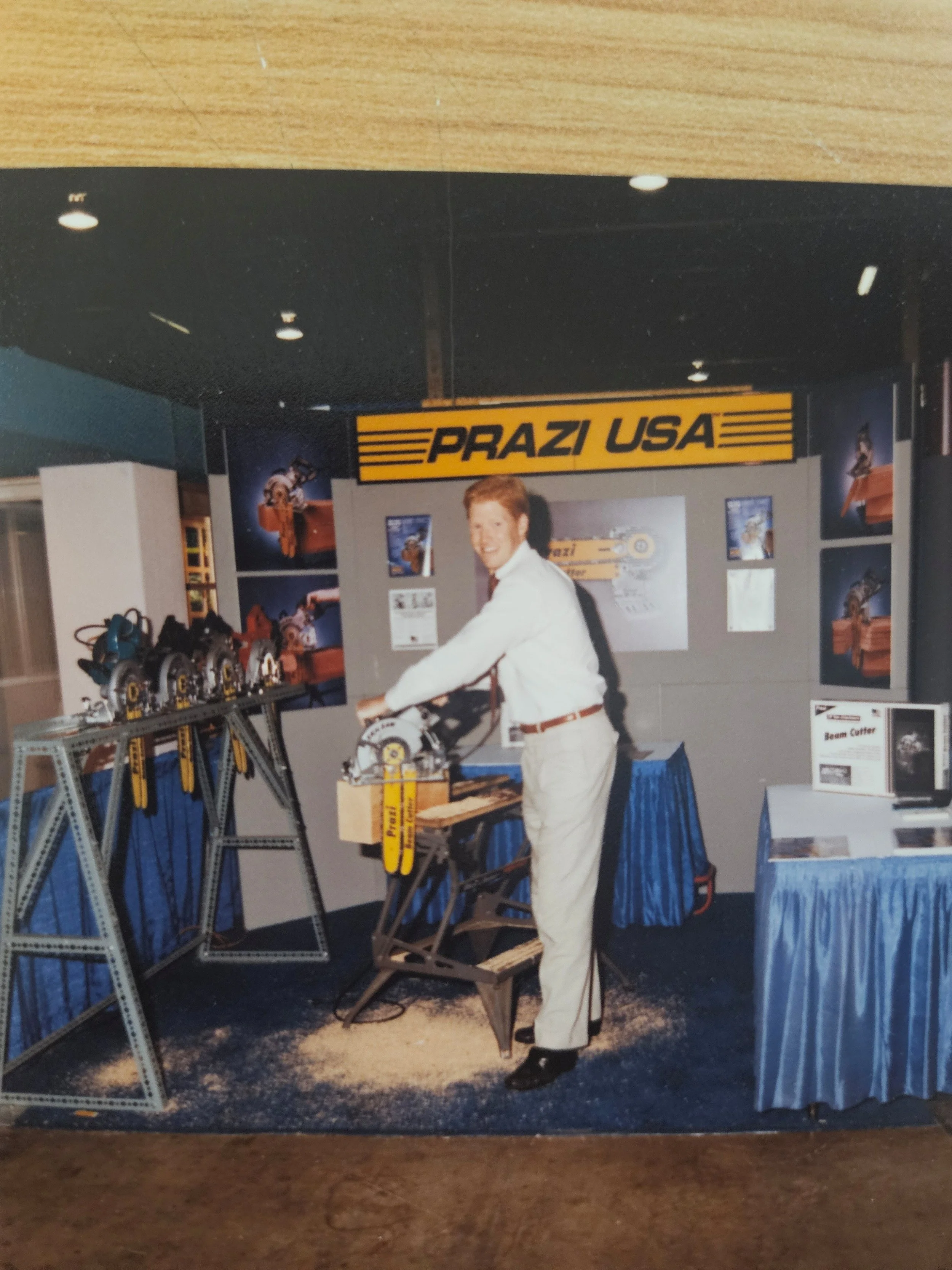 A man in white suits demonstrates a power saw at a trade show booth with a yellow 'PRAZI USA' sign, various machinery displays, and informational posters.