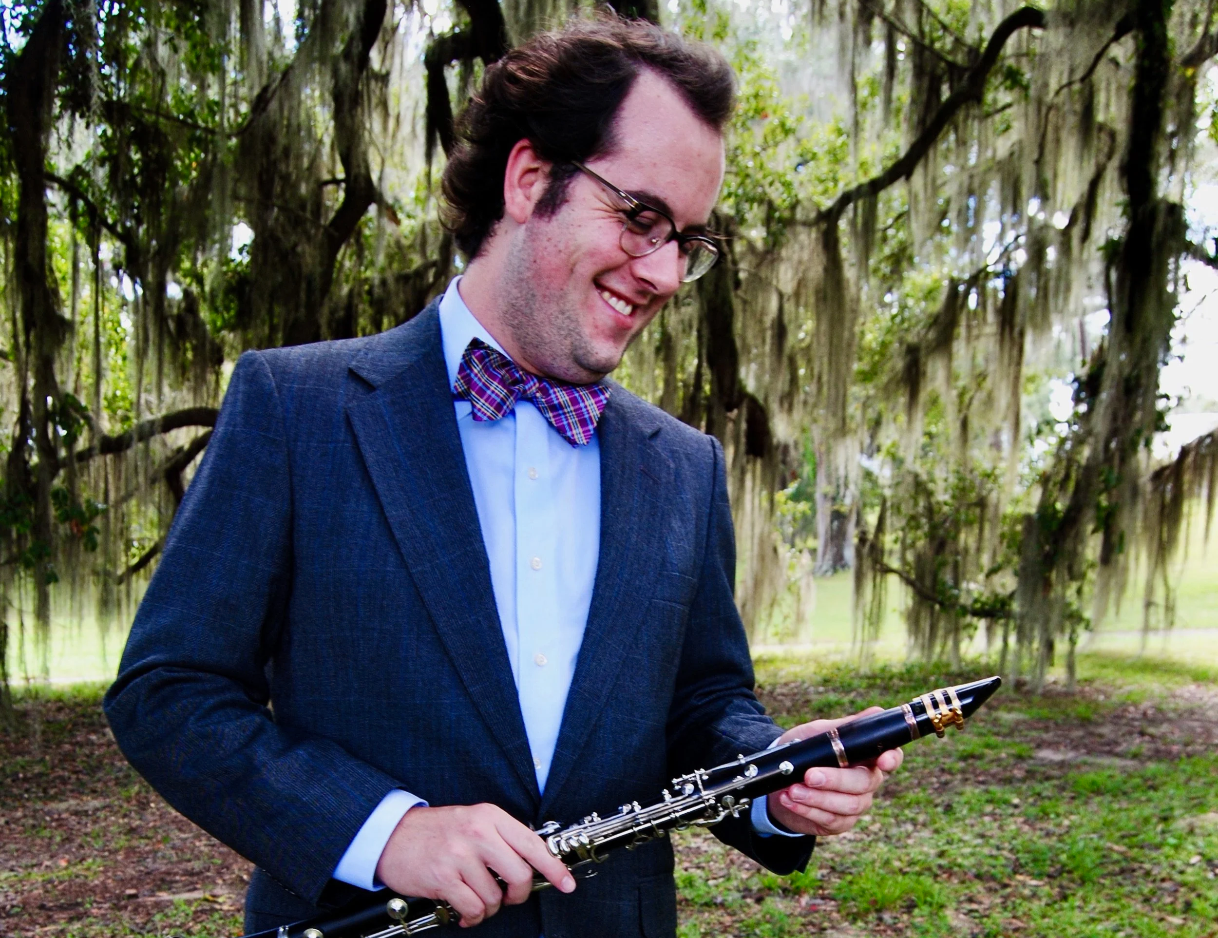 Connor in a blue suit and bow tie smiling while holding a clarinet outdoors with trees and Spanish moss hanging from branches in the background.