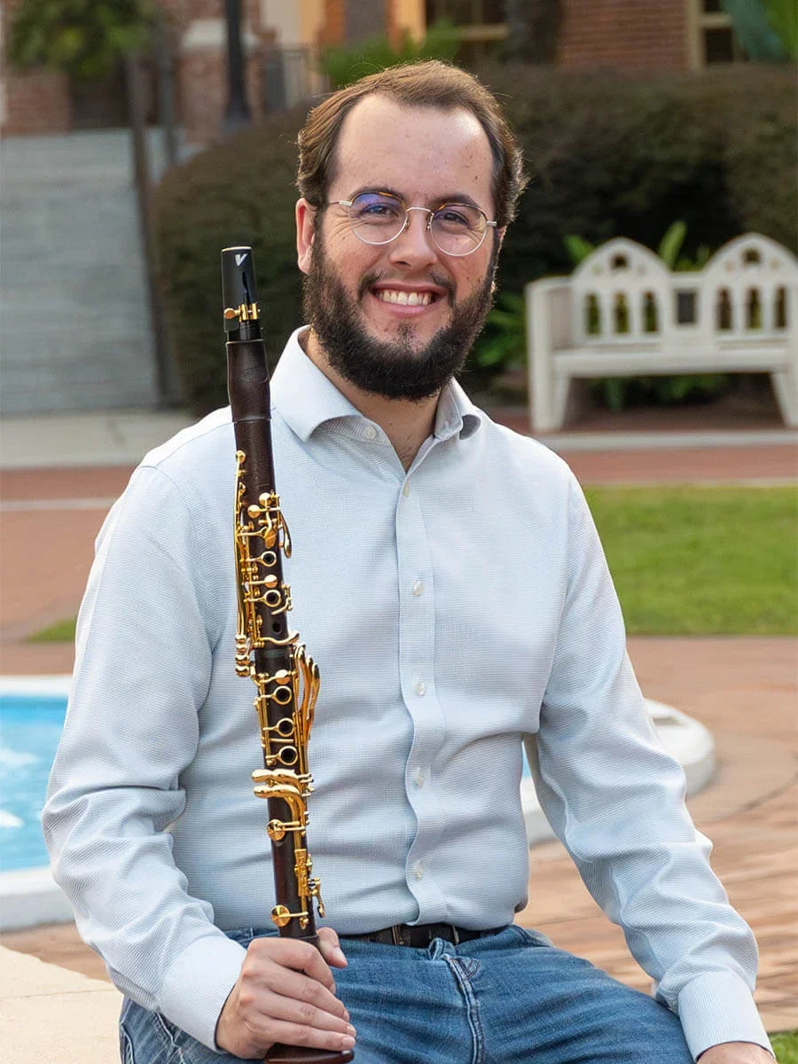 Connor, holding a clarinet, smiling at the camera, with a building, benches, and greenery in the background.