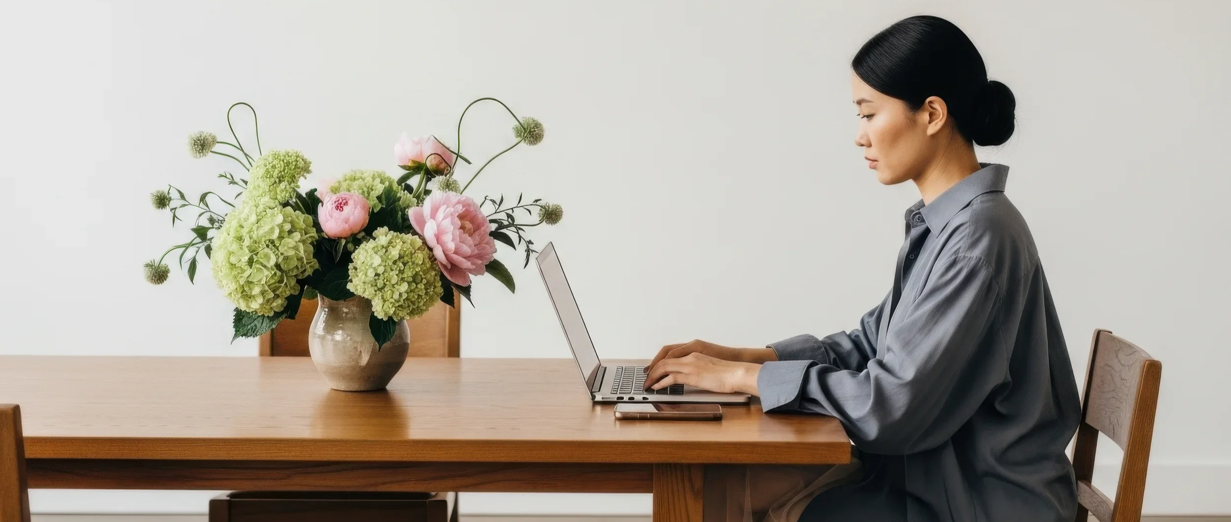 Woman sitting at a wooden table working on a laptop, with a bouquet of pink and green flowers in a ceramic vase on the table.