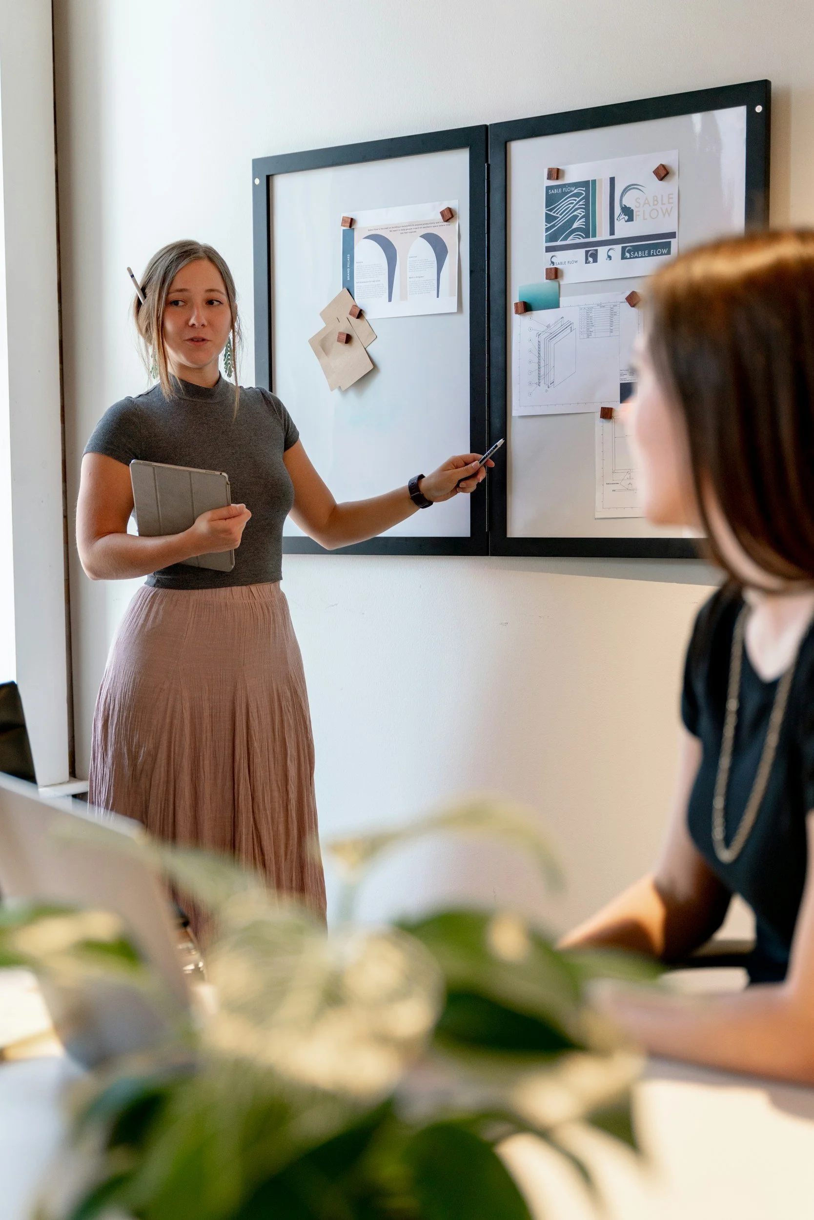 A woman presenting to another woman in a meeting room with a whiteboard filled with papers and notes.