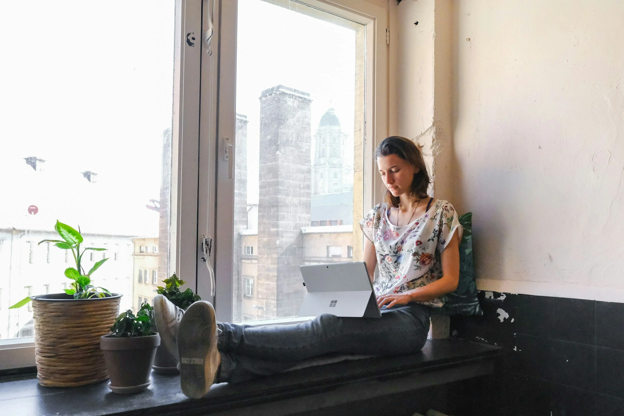 A young woman sitting on a windowsill inside a building, working on a tablet. There are potted plants nearby, and tall buildings can be seen through the window.