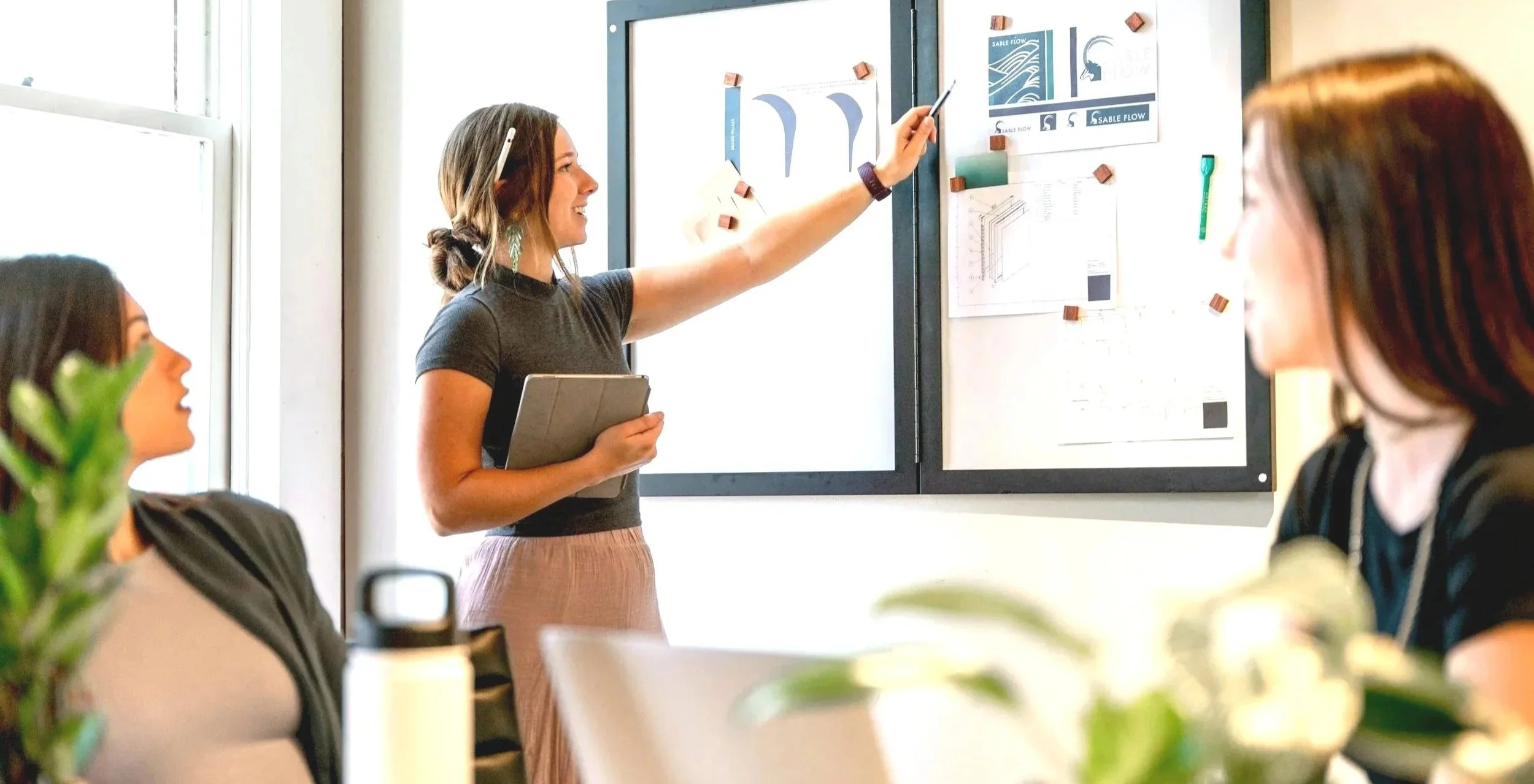 A woman presents to a group of three women seated at a table, with a large whiteboard filled with charts and diagrams behind her, in a bright conference room.