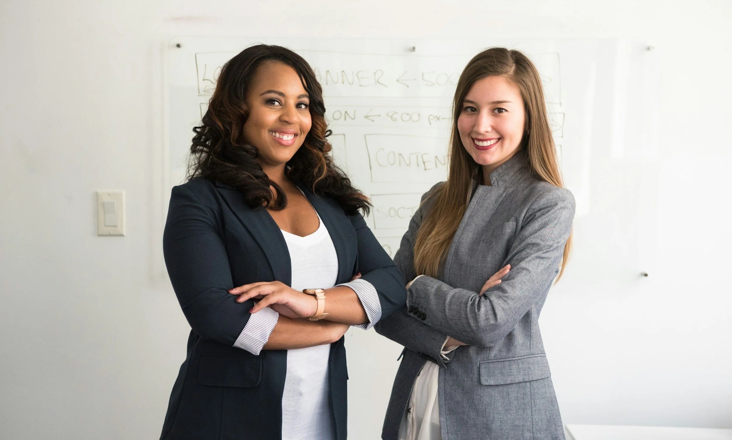 Two women in business attire standing close together and smiling in front of a whiteboard in a professional setting.