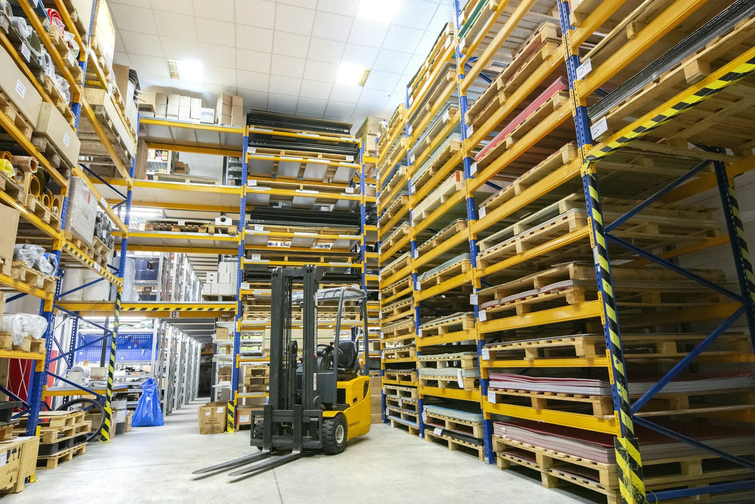 A warehouse with tall yellow and blue shelving units stocked with various items, a yellow forklift in the center, and concrete flooring.