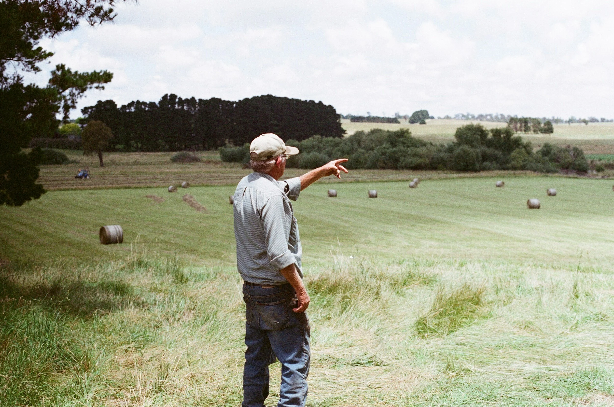 A man in a grey shirt and cap standing on a grassy hill, pointing towards a field with hay bales, trees, and a partly cloudy sky.