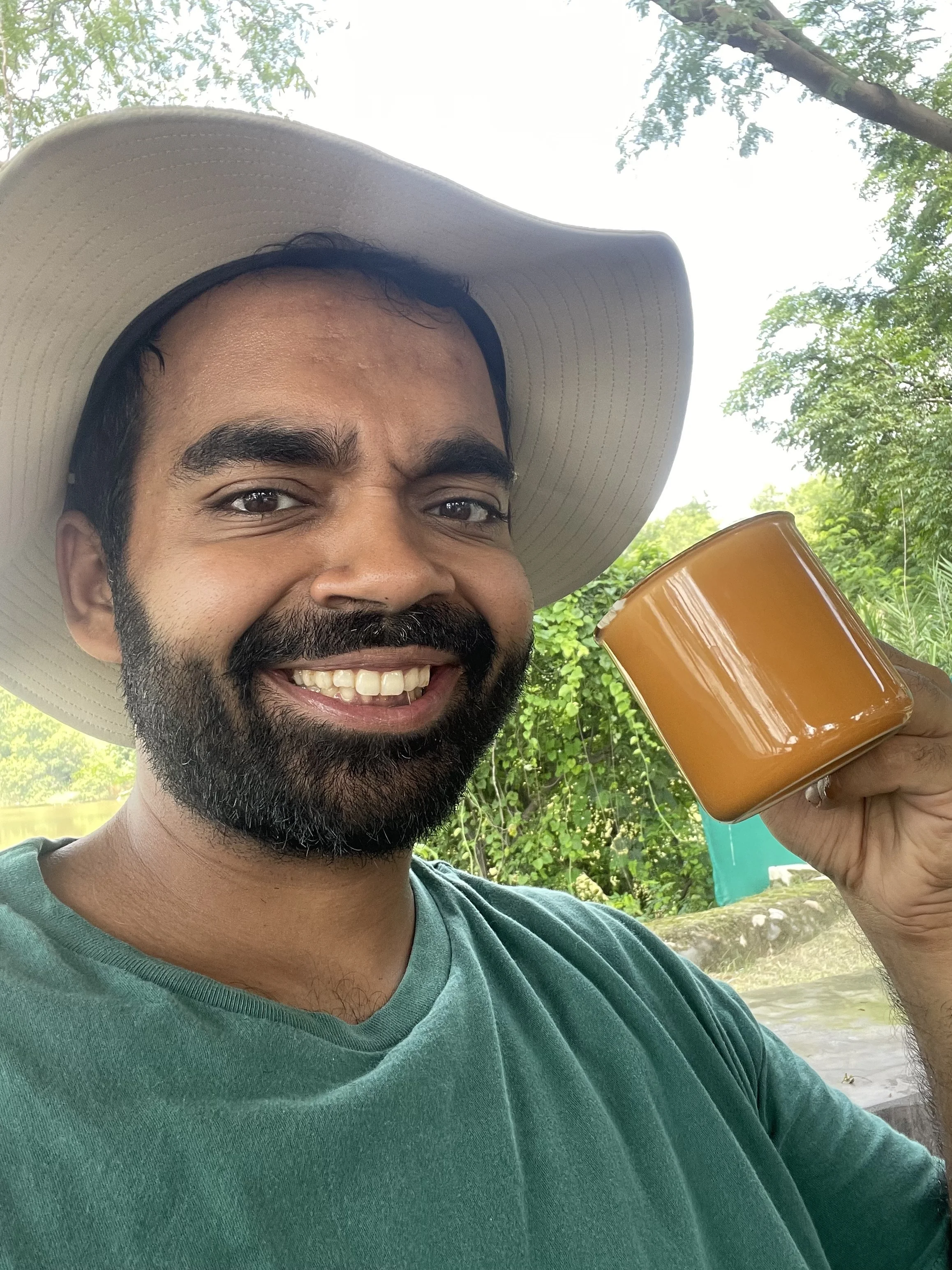 A man in a large beige sun hat smiling and holding a pot of brown beverage outdoors with green trees in the background.