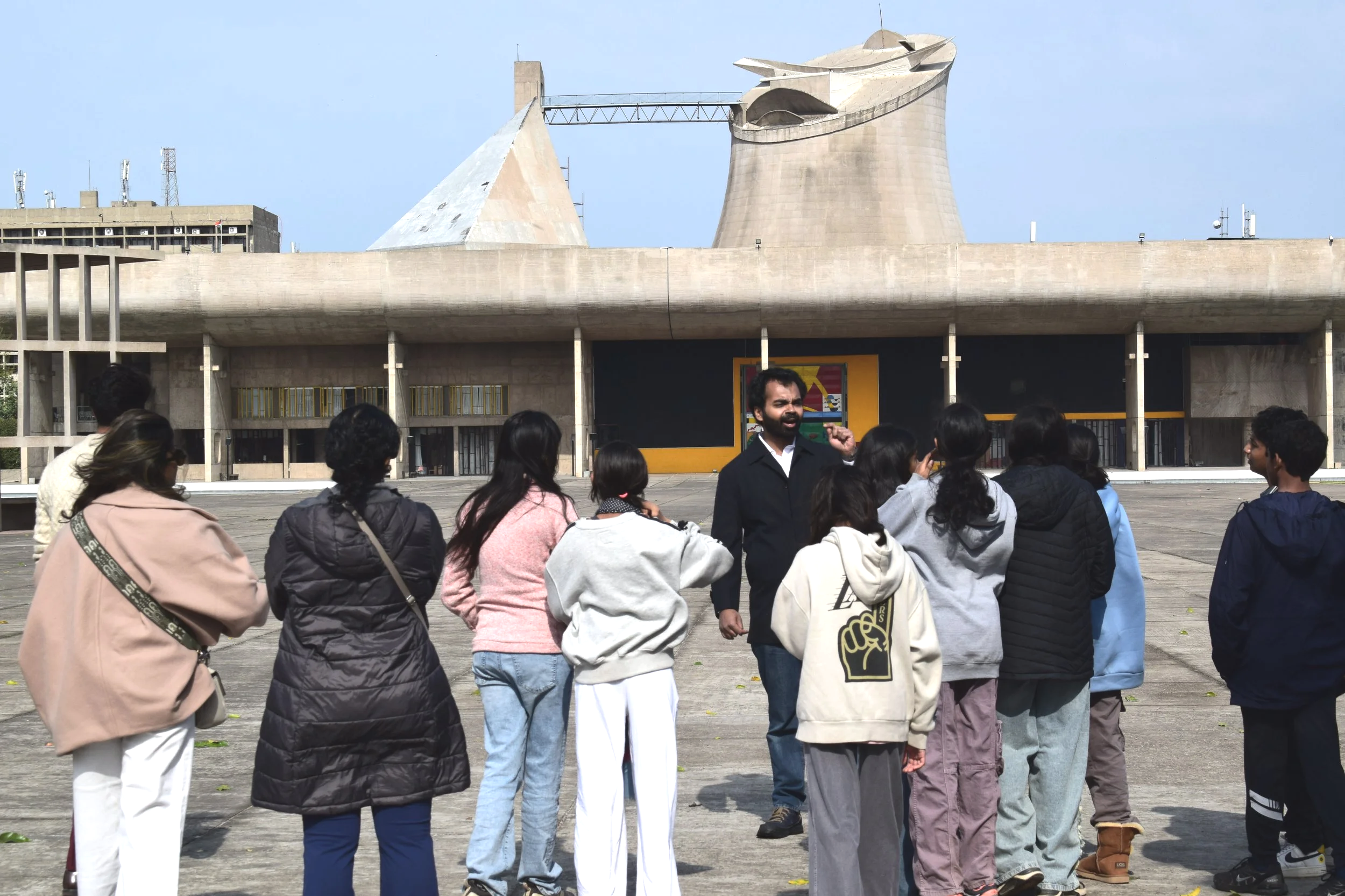 A group of people, including men and women, standing outdoors in front of a large concrete building with unique architecture, including sculptures on top, on a clear day.