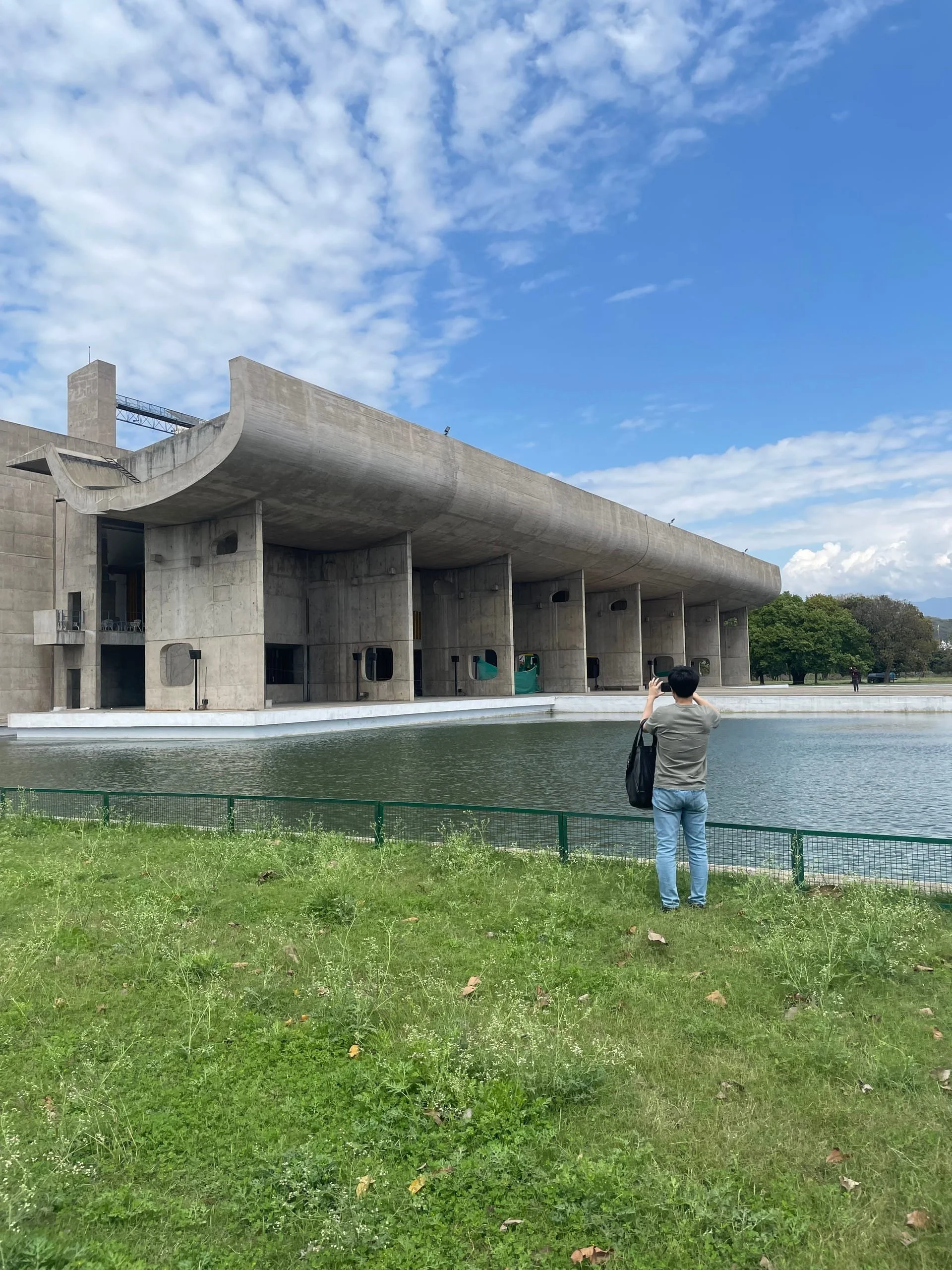 A person wearing a gray shirt and jeans is standing on green grass, taking a photo of a large concrete building with water in the foreground and trees in the background under a partly cloudy sky.