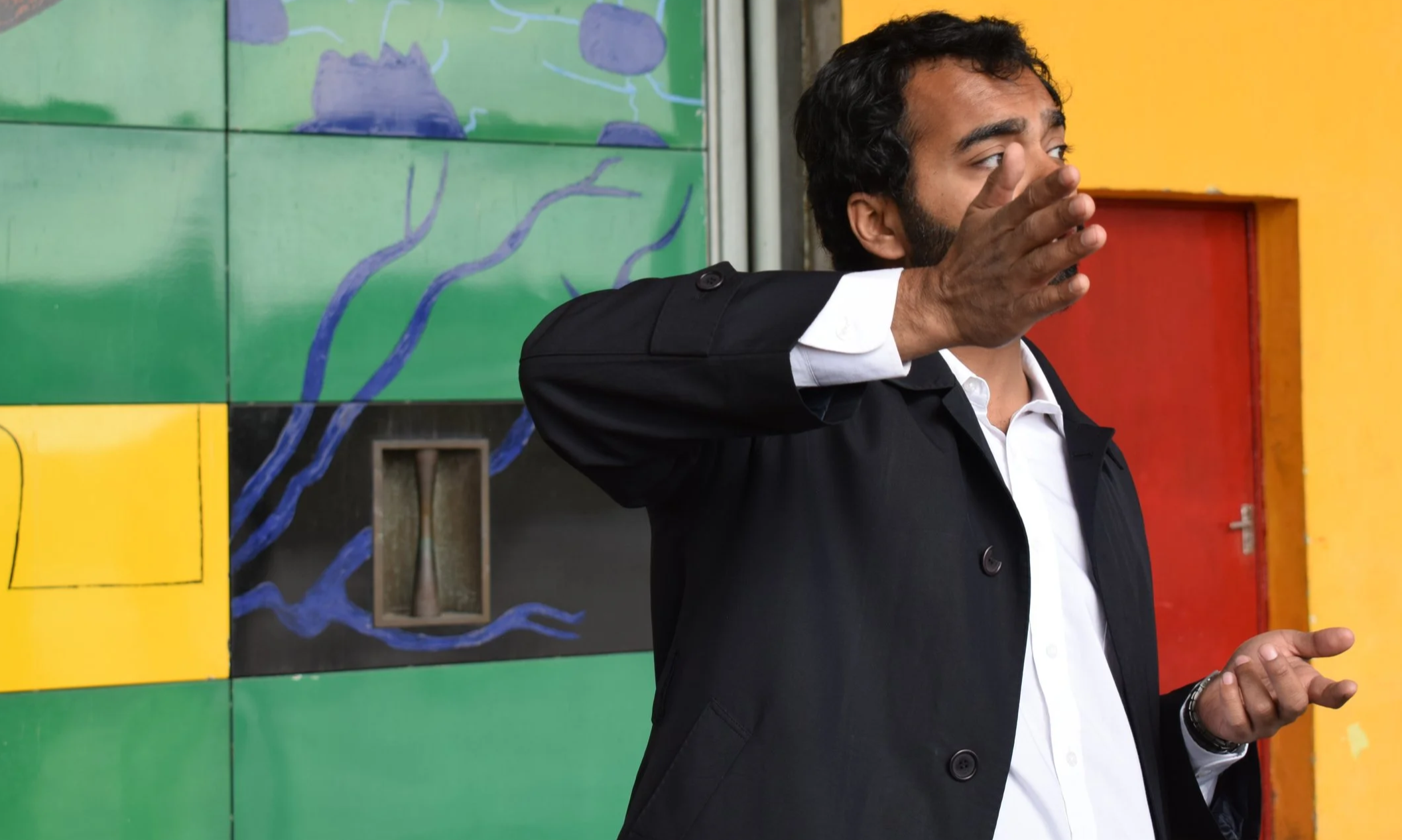 A man in a black suit with a white shirt is gesturing with his right hand while speaking against a colorful background with green, yellow, and red panels and a mural featuring blue lines and shapes.