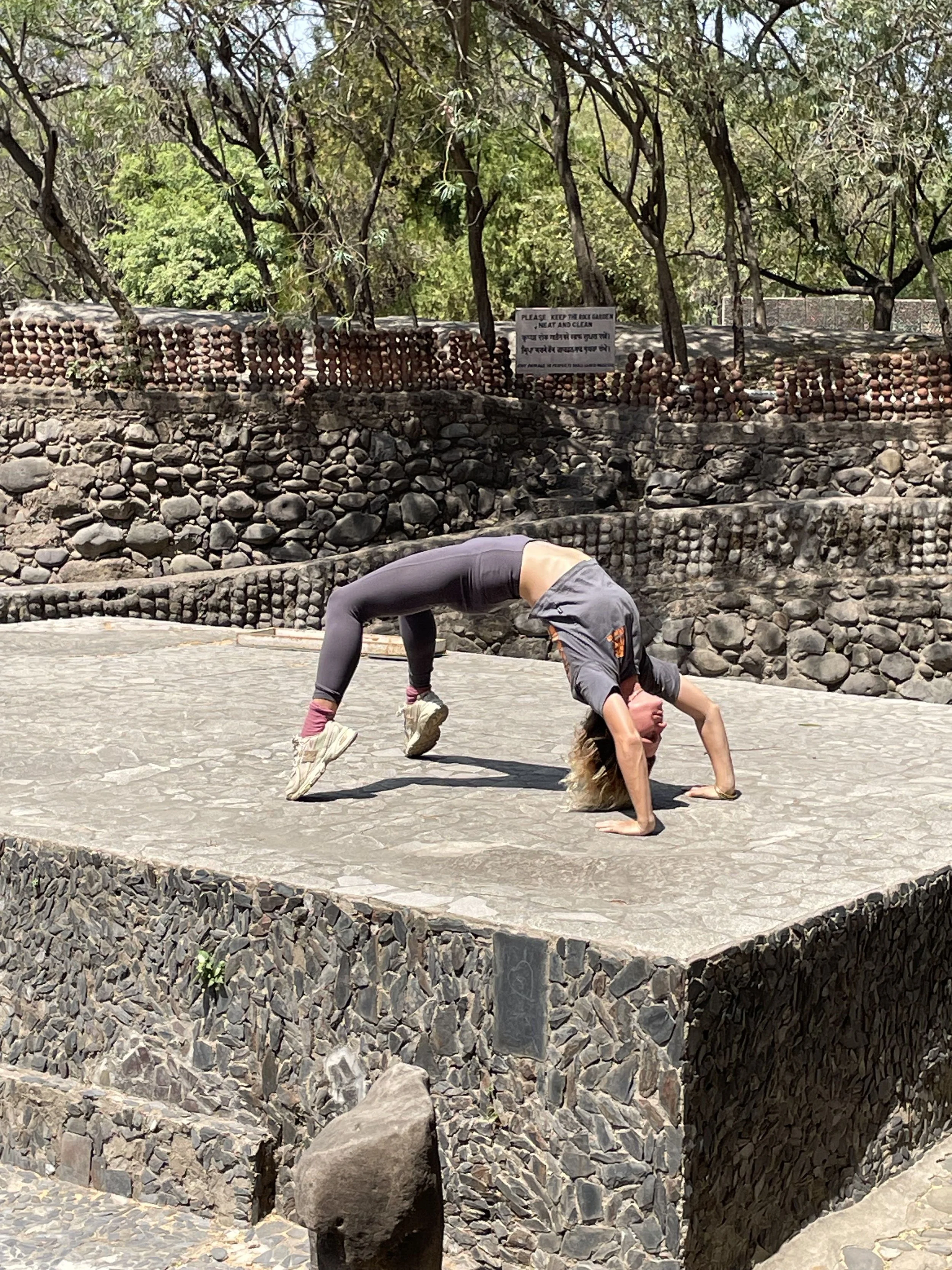 A person performing yoga outdoors on a stone platform with trees in the background. The person is in a backbend pose, with hands and feet on the ground and back arched upward.