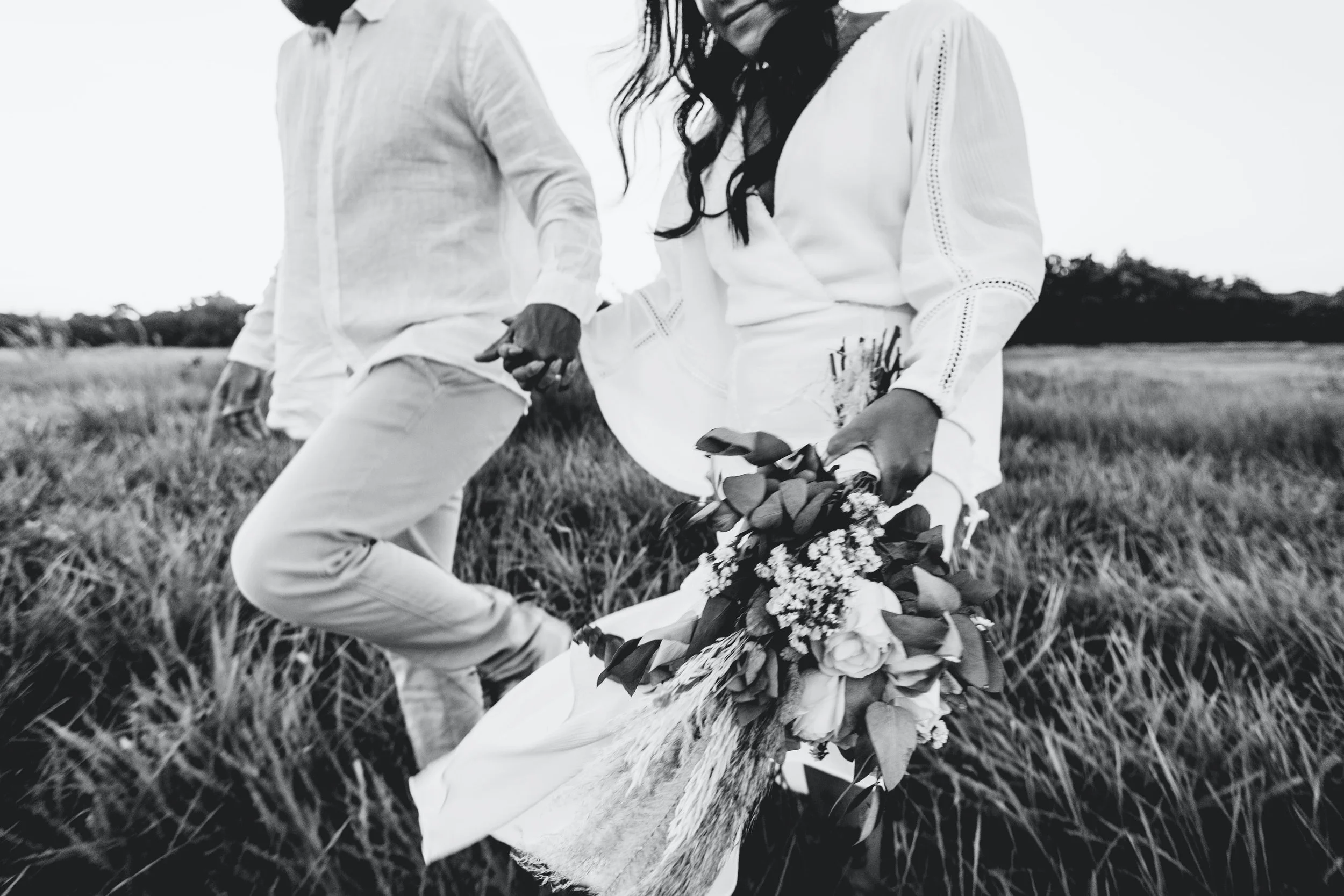 A black and white photo of a couple holding hands in a grassy field. The woman is holding a bouquet of flowers, and they are dressed in light-colored clothing.