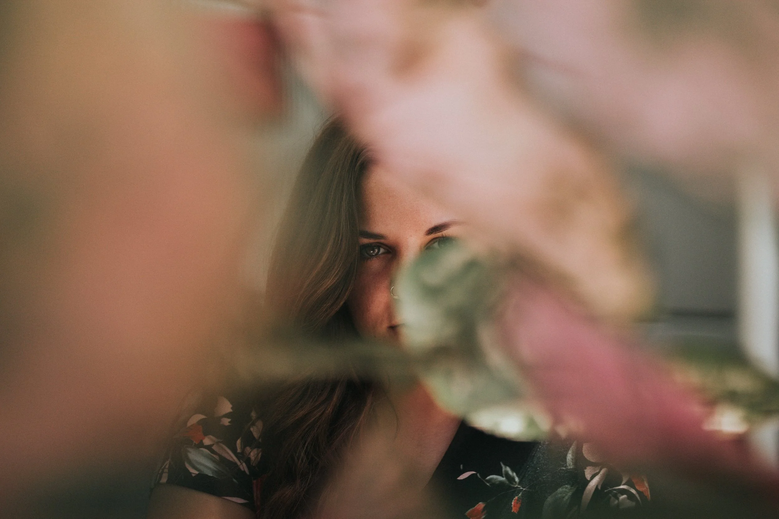 A woman with long brown hair and a nose ring, partially obscured by colorful flowers, looking directly at the camera.