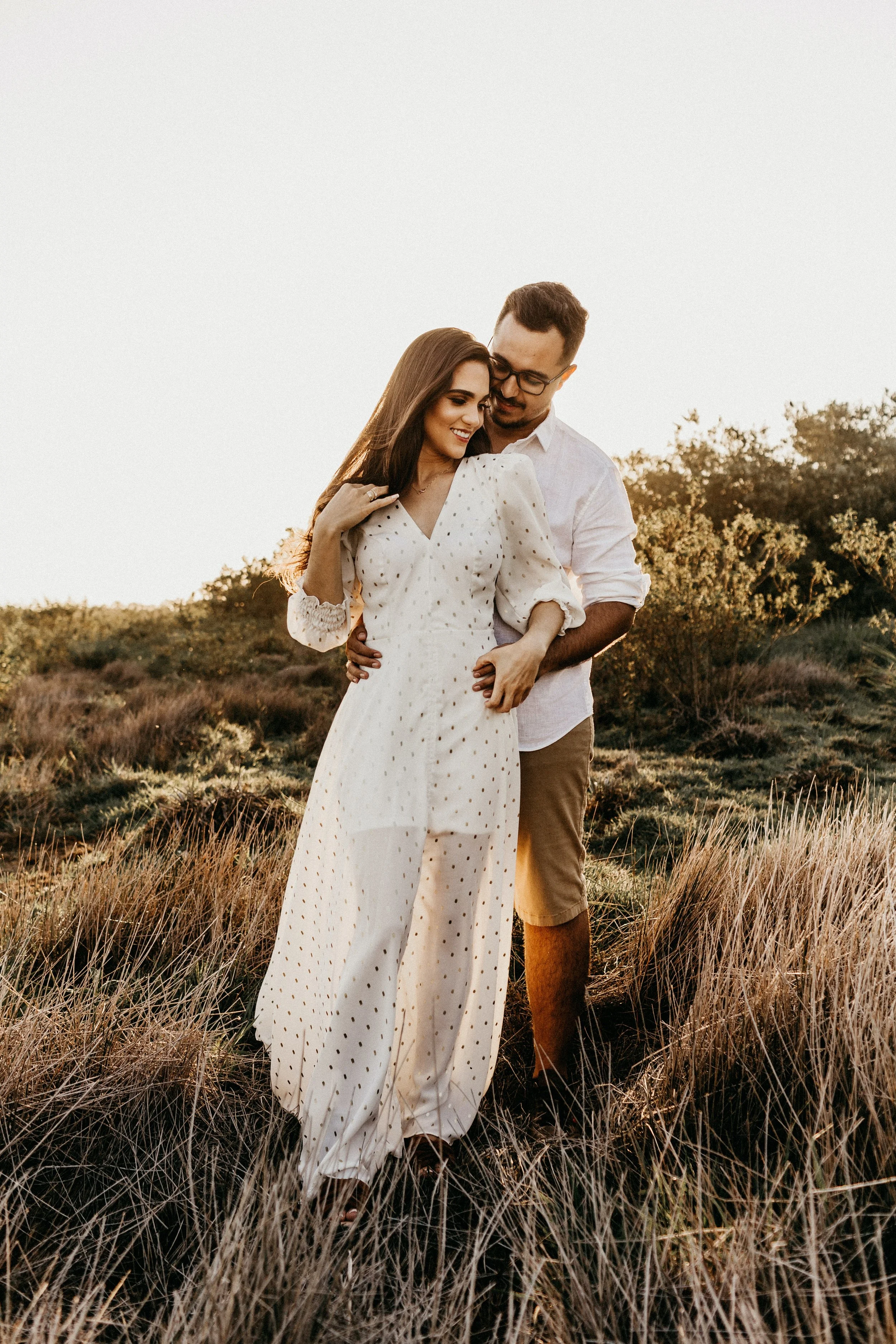 A couple standing close together in a field of tall grass and bushes, with the woman in a white polka-dot dress and the man in a white shirt and beige shorts, during sunset.