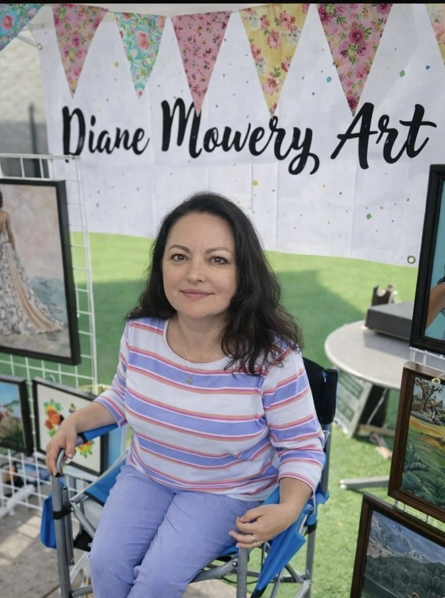 A woman sitting in a blue wheelchair at an art booth with paintings on display, a sign that reads 'Diane Mowery Art,' and pastel-colored bunting hanging above.