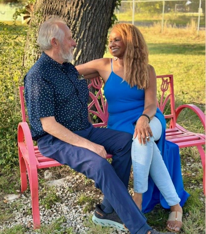 A man and woman sit on a pink park bench outdoors, smiling and looking at each other. The woman has her arm around the man."