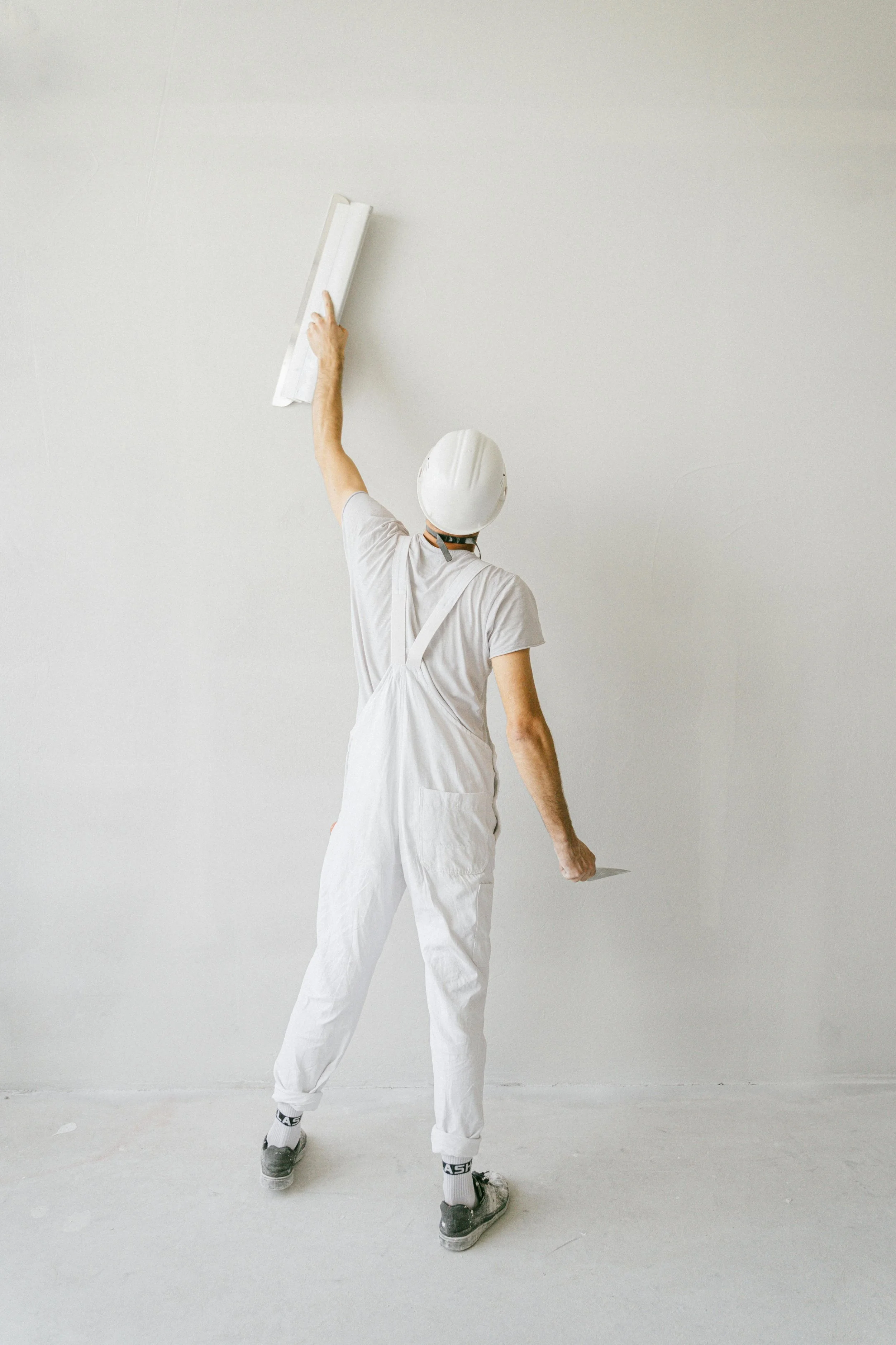 A person dressed in white overalls and a white helmet is painting a wall with a roller brush.