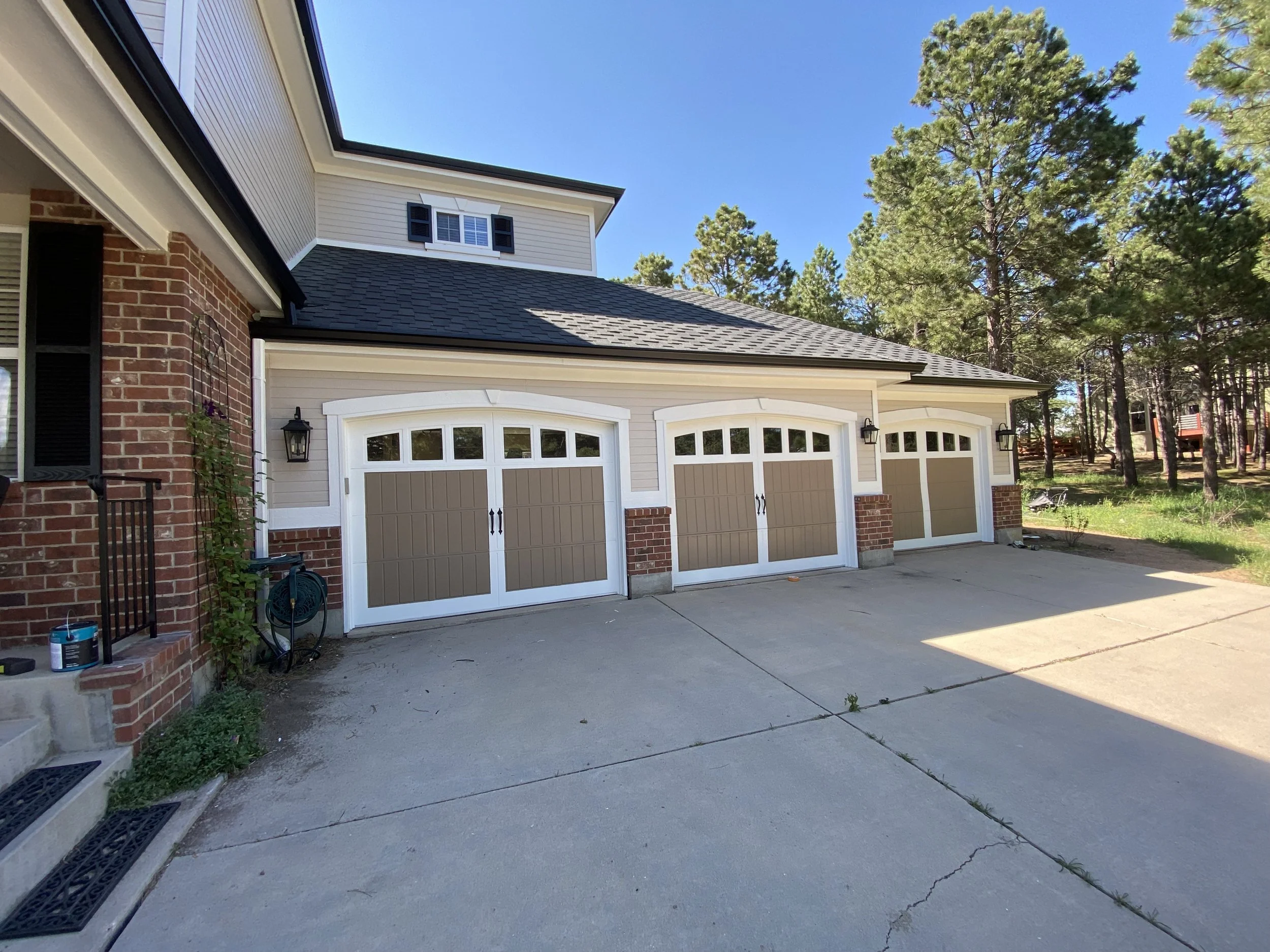 Three car garage with white and tan doors, brick and siding exterior, black lantern style lights, concrete driveway, and wooded area with tall trees in the background.