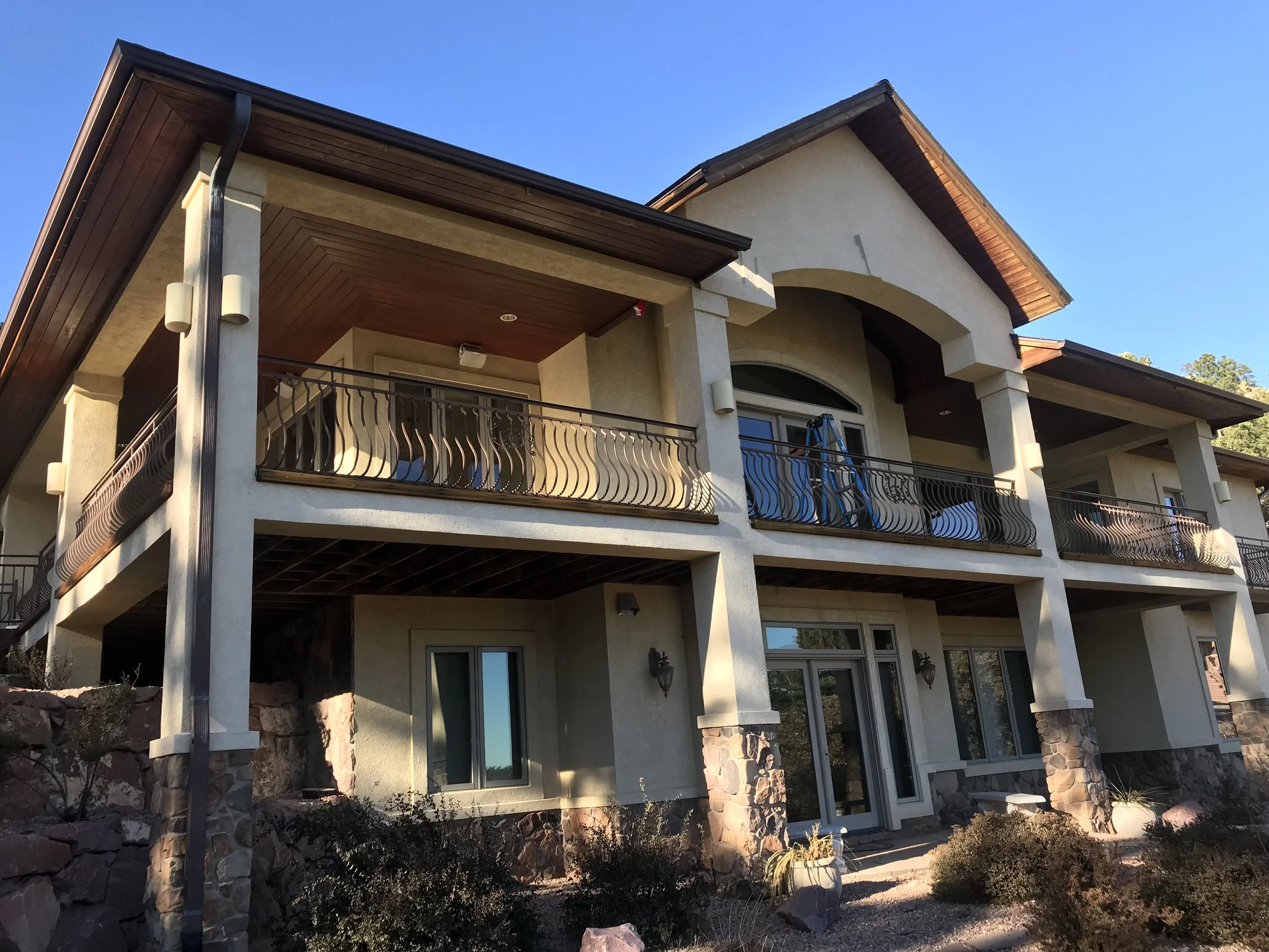 Two-story modern house with beige stucco exterior, stone accents at the base, and a large balcony with metal railings on the upper level. The house features multiple windows, a wooden ceiling on the balcony, and is set against a clear blue sky.