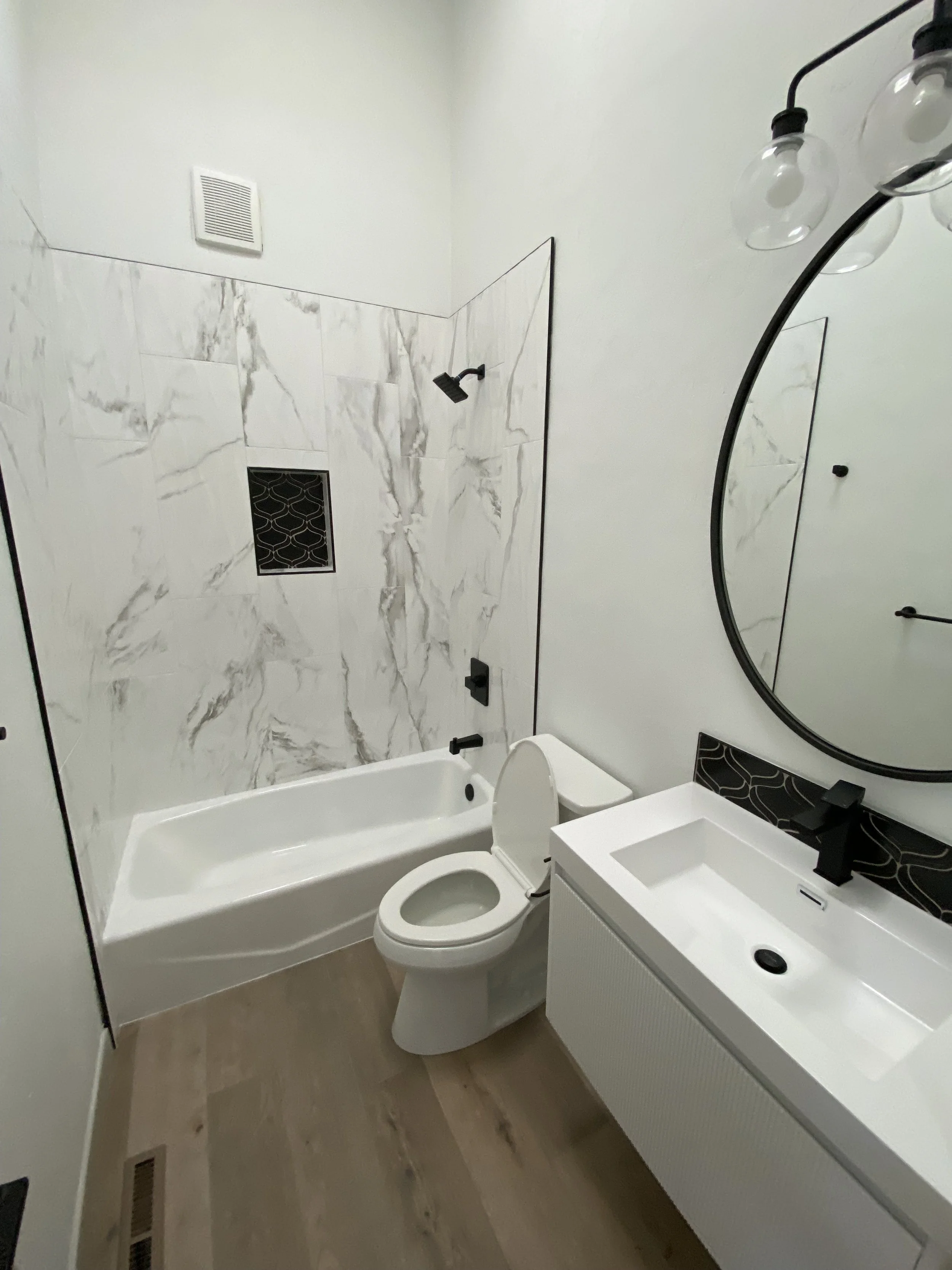 Modern bathroom with white walls, marble shower wall, bathtub, toilet, white sink vanity, round mirror, black fixtures, and clear light bulbs.