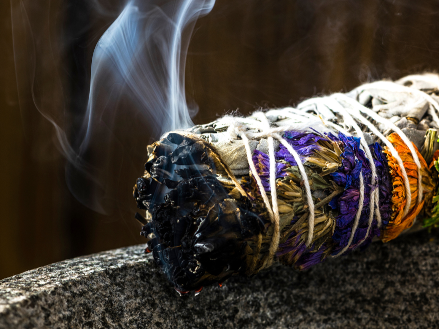 A smoldering sage bundle with smoke rising from burnt herbs, placed on a stone surface.