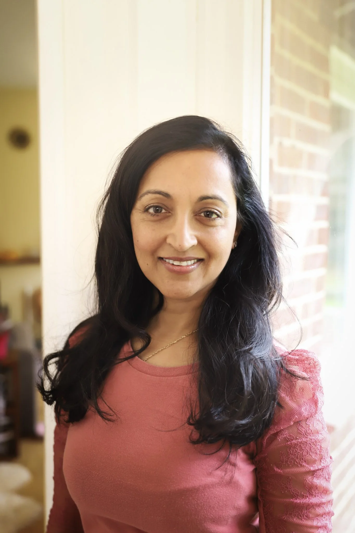 Roxana with long, dark hair smiling at the camera inside a home near a window with a brick wall outside.