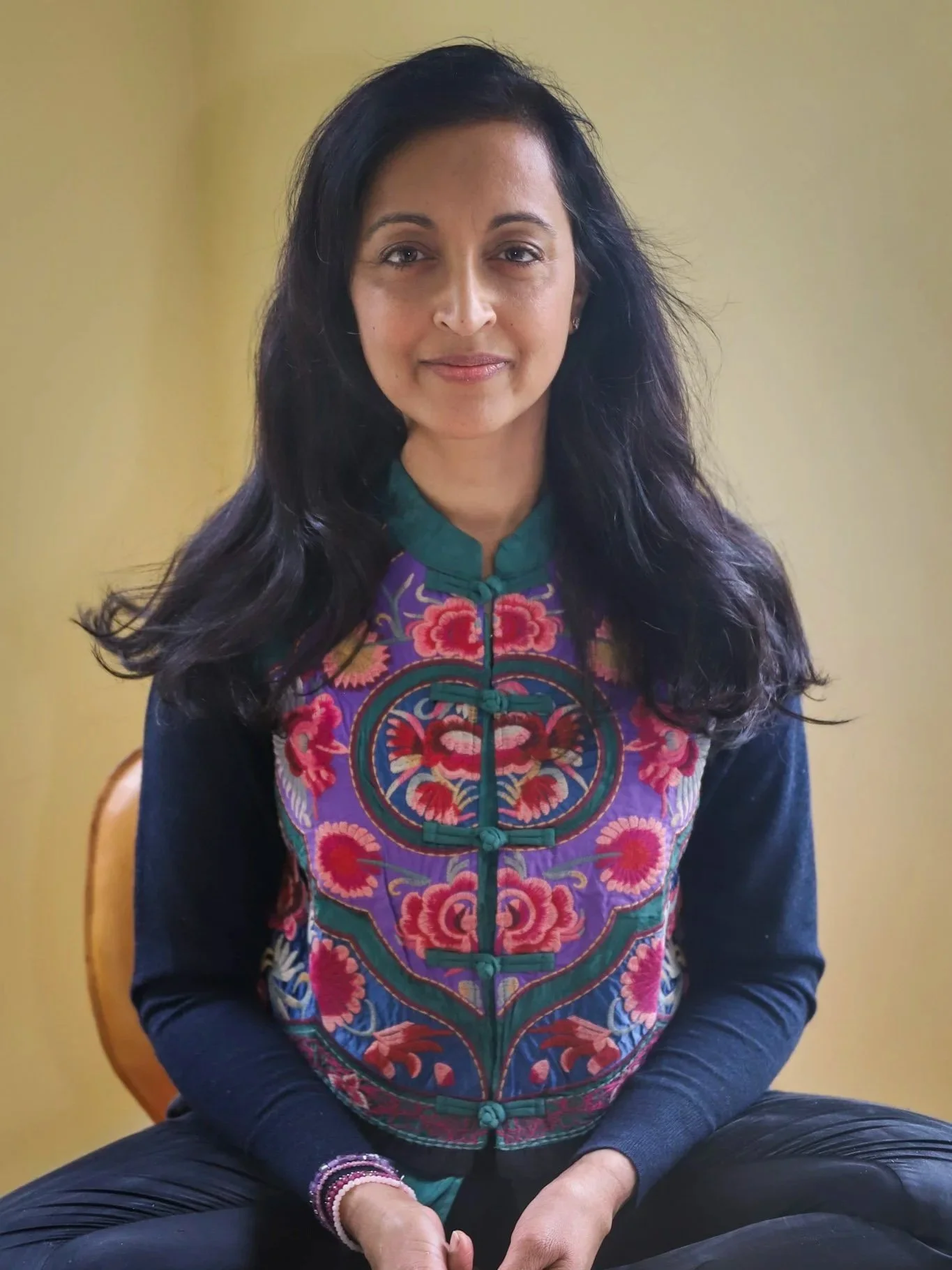 Roxana with dark hair wearing a colorful embroidered top sitting against a plain beige wall.